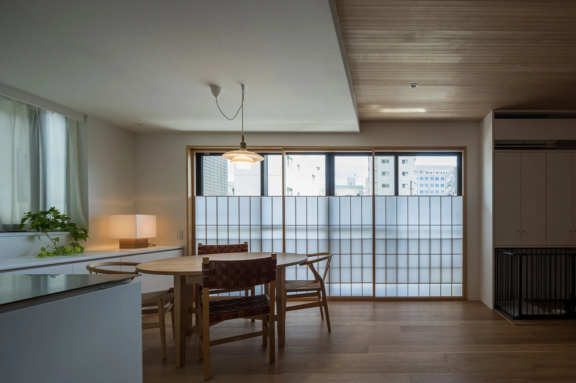 A minimalist dining room features an oval wood table with woven chairs in front of sliding shoji screens under a wood-paneled ceiling.