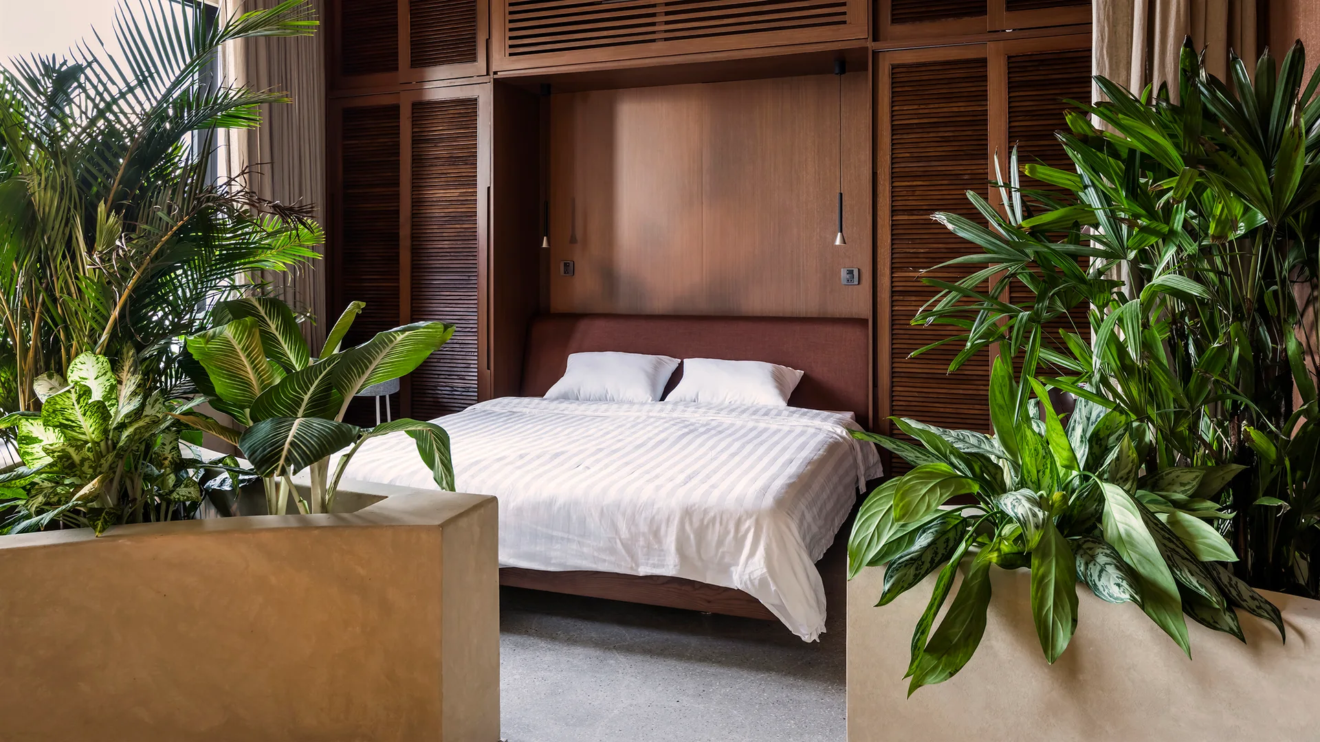 A bedroom featuring a bed set against dark wood louvered cabinets, flanked by two large concrete planters filled with lush green houseplants.