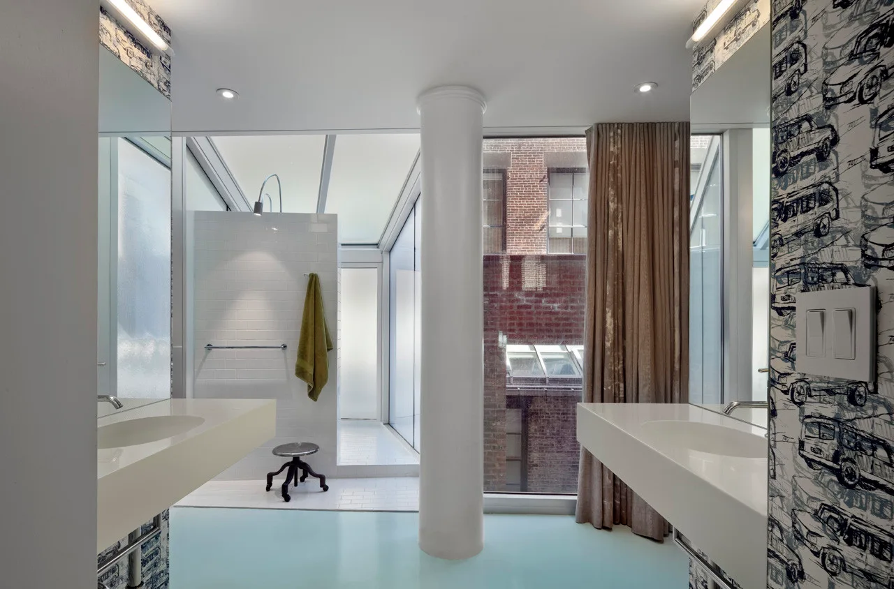A modern bathroom features two white floating vanities facing each other, a central structural column, and a large window looking out to a brick facade.