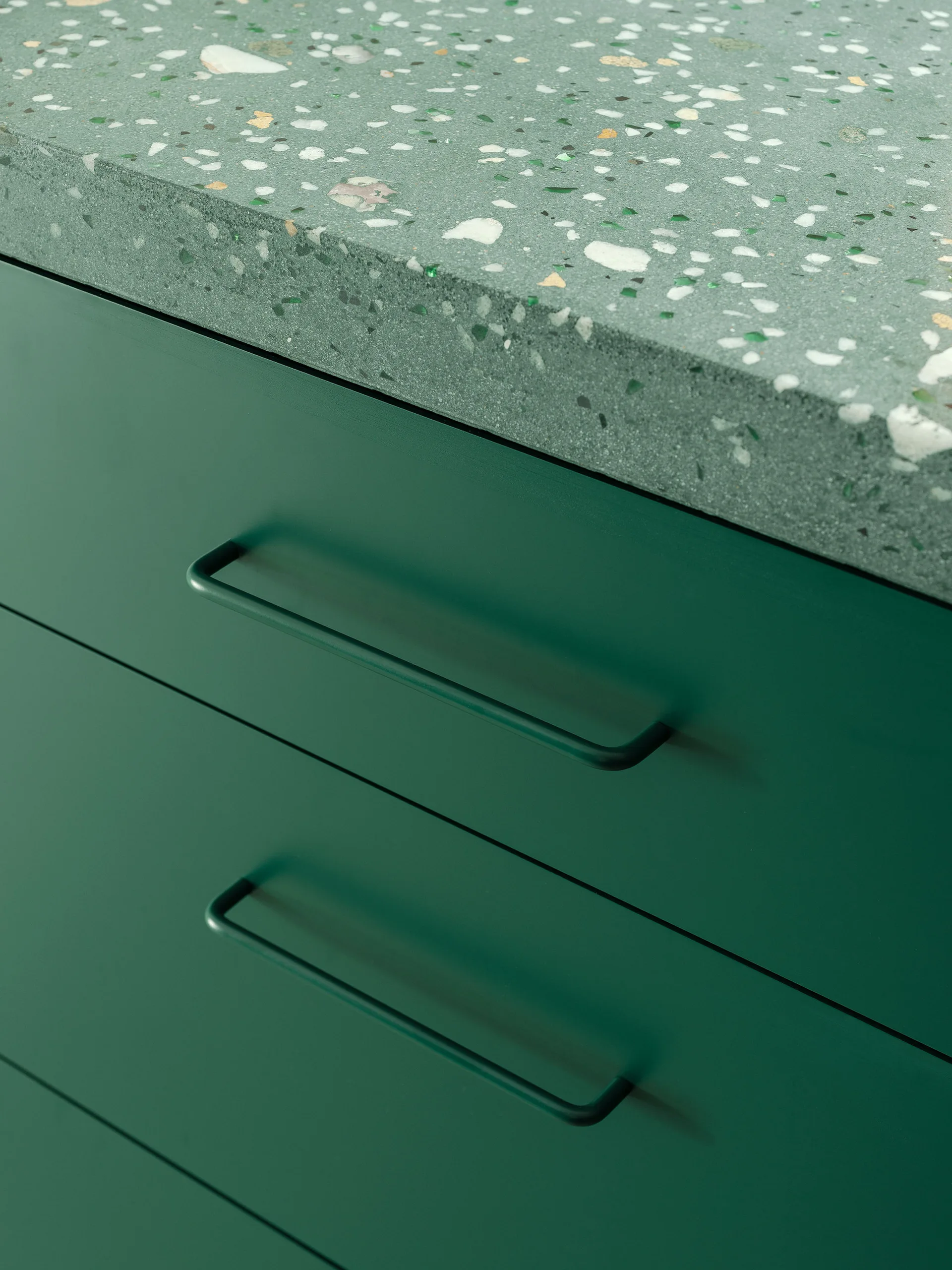A close-up of matte green kitchen cabinets with thin matching handles beneath a green terrazzo countertop with light speckled chips.