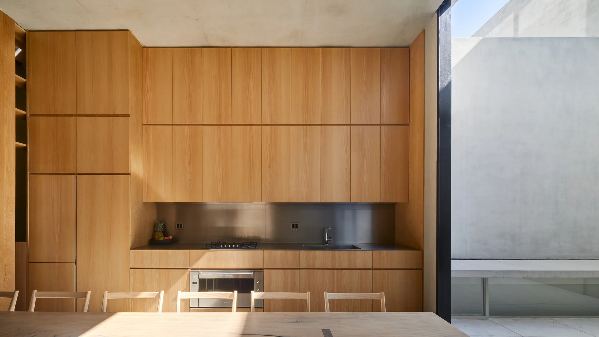 A minimalist kitchen features full-height wood cabinetry, a stainless steel countertop and backsplash, and a wooden dining table in the foreground.