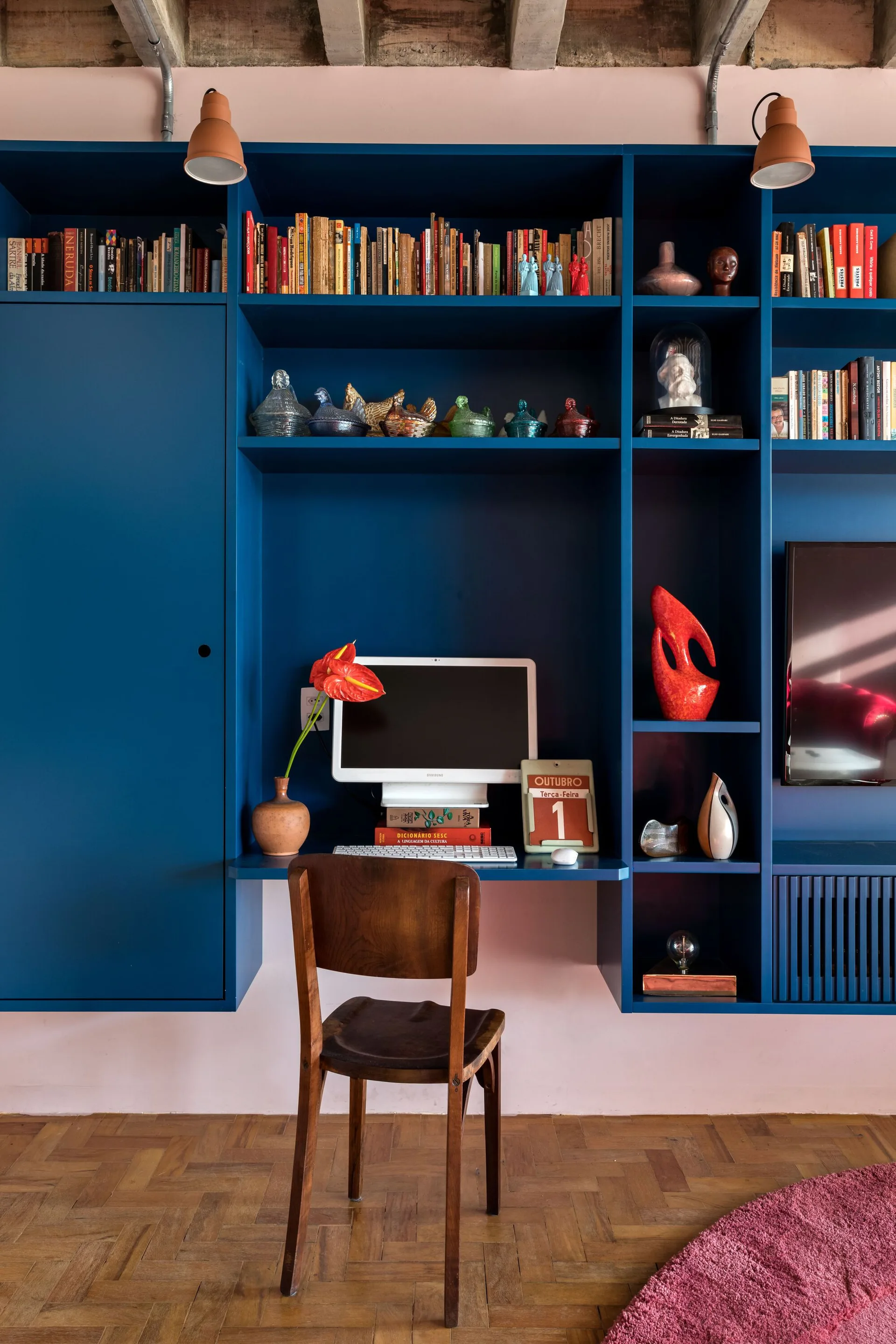 A blue built-in shelving unit with an integrated desk and computer sits on parquet wood flooring in front of a light pink wall.