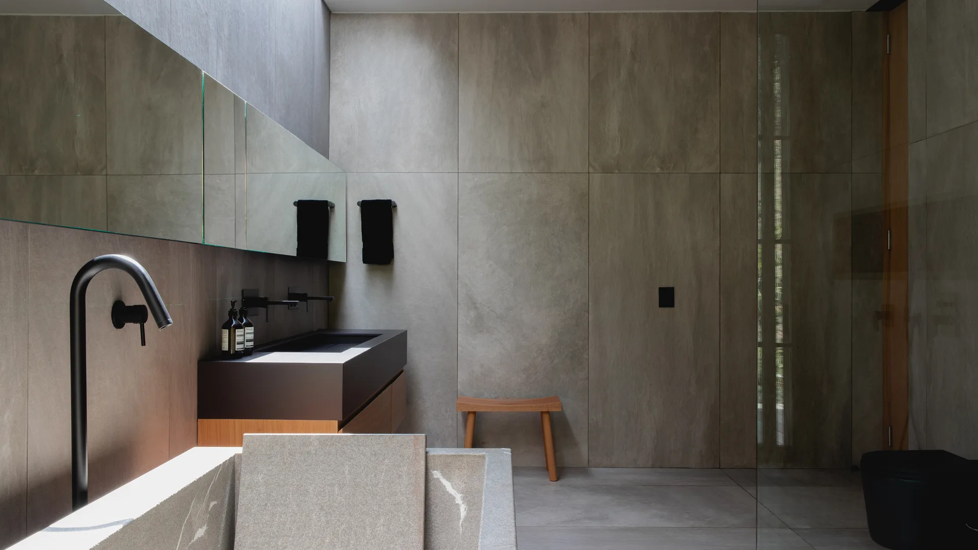 A minimalist bathroom features large grey stone wall tiles, a dark floating vanity with a wooden base, a skylight, and a small wooden stool.