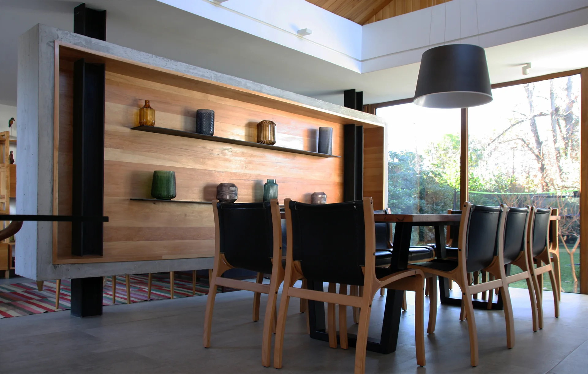 A modern dining room features a concrete partition wall lined with wood paneling and shelving next to a wooden dining set.