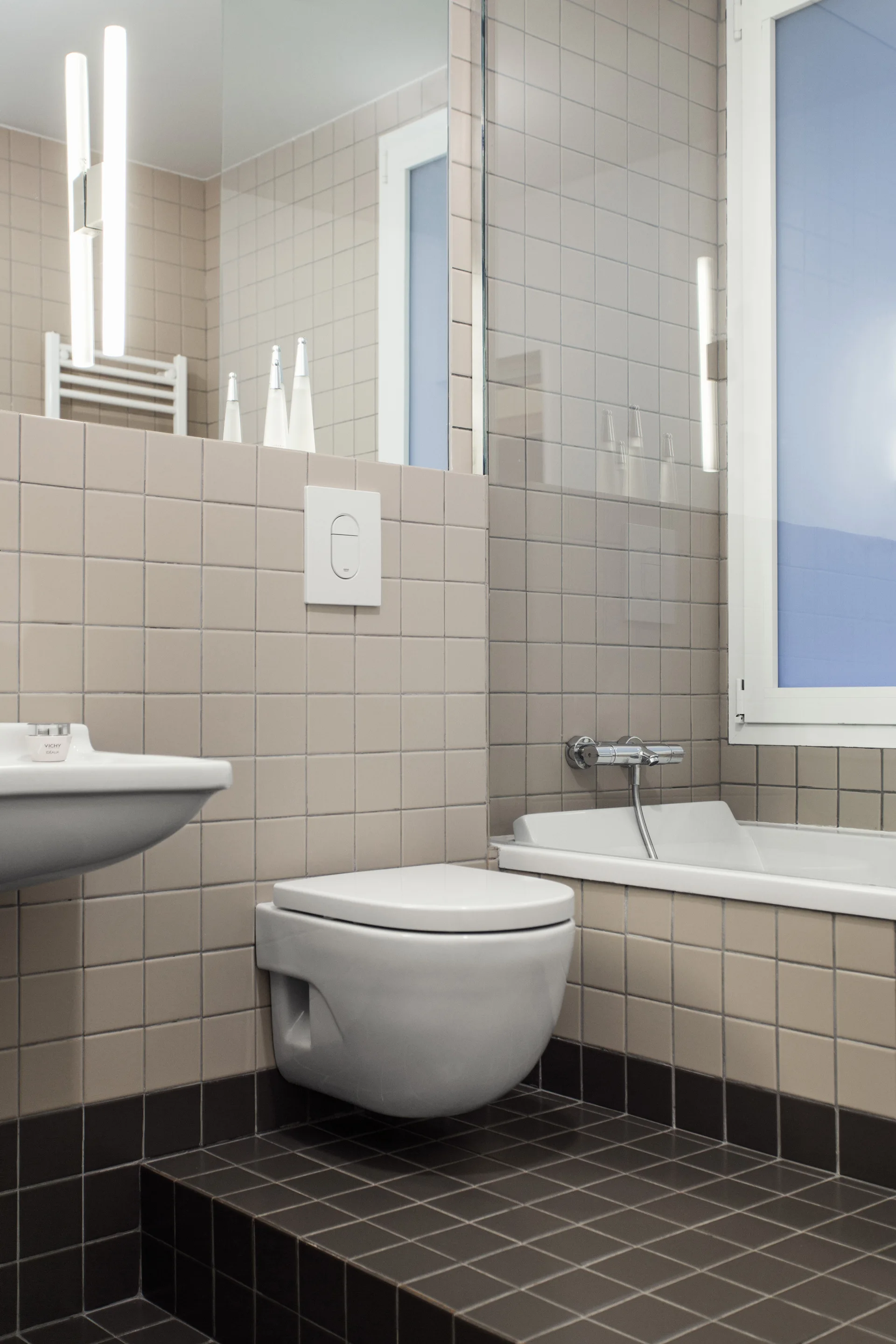 A contemporary bathroom features a wall-mounted white toilet, a built-in bathtub, and beige square wall tiles contrasted with dark brown floor tiles.