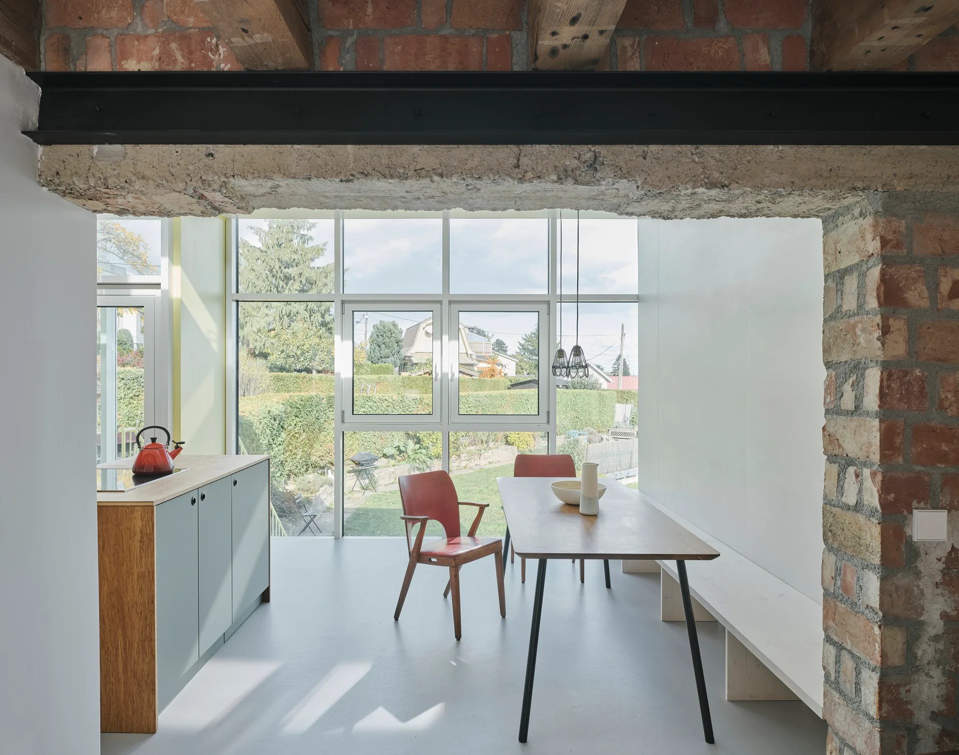A minimalist dining area featuring a wooden table, built-in bench, and kitchen cabinets adjacent to exposed brick walls and garden-facing windows.
