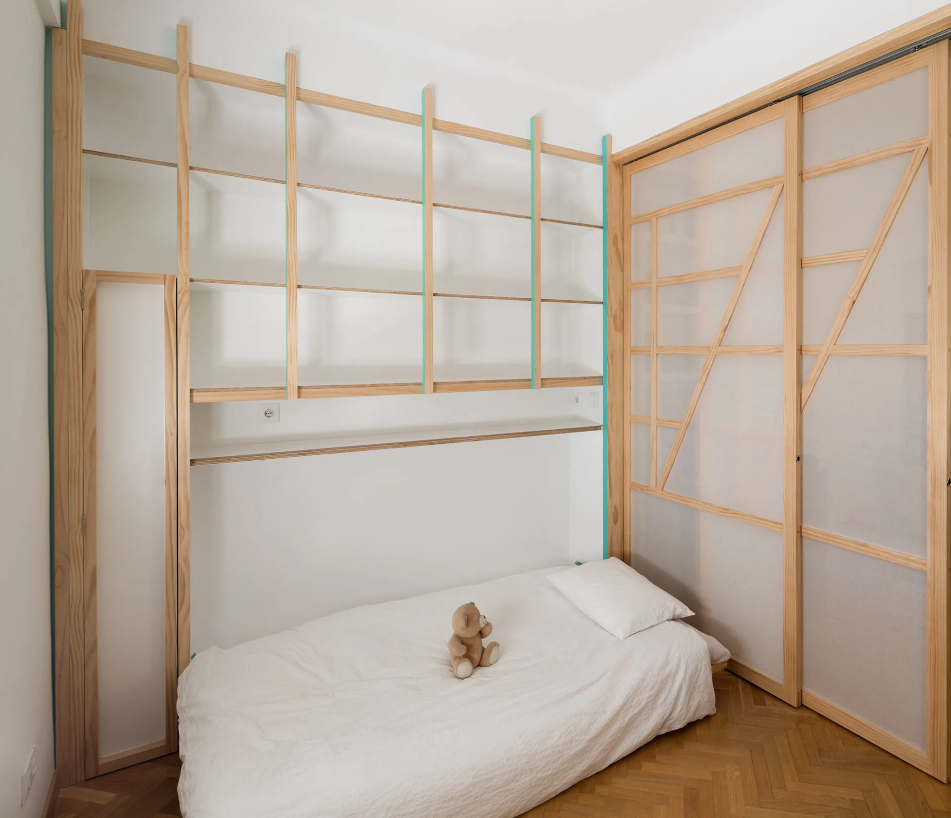 A minimalist bedroom featuring a low white bed, herringbone parquet floors, and custom light wood shelving with sliding screen doors.