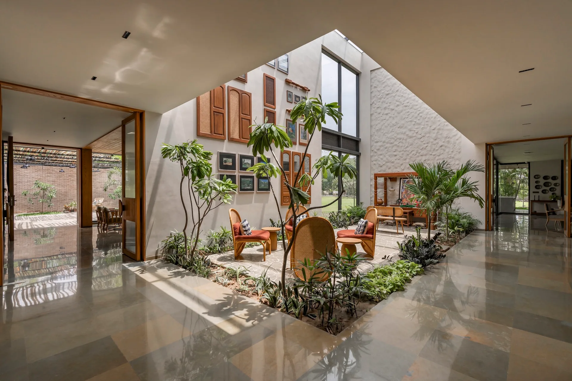 An indoor courtyard atrium features rattan armchairs, a central garden bed with trees, and a gallery wall with wooden shutters beneath a high skylight.