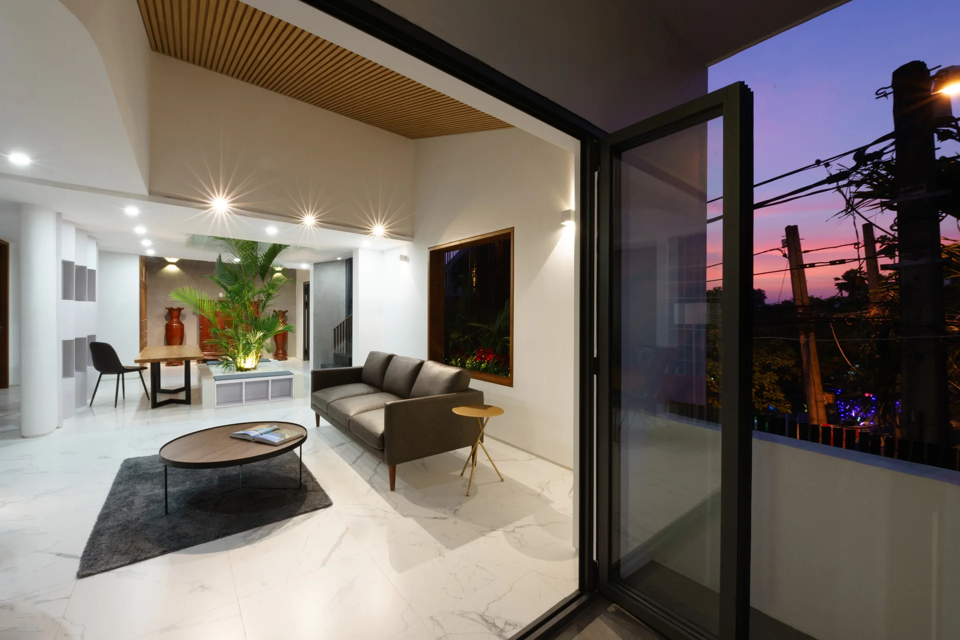 A modern living room features a dark grey sofa, a slatted wood ceiling, marble-patterned floor tiles, and a large open glass door showcasing a sunset.