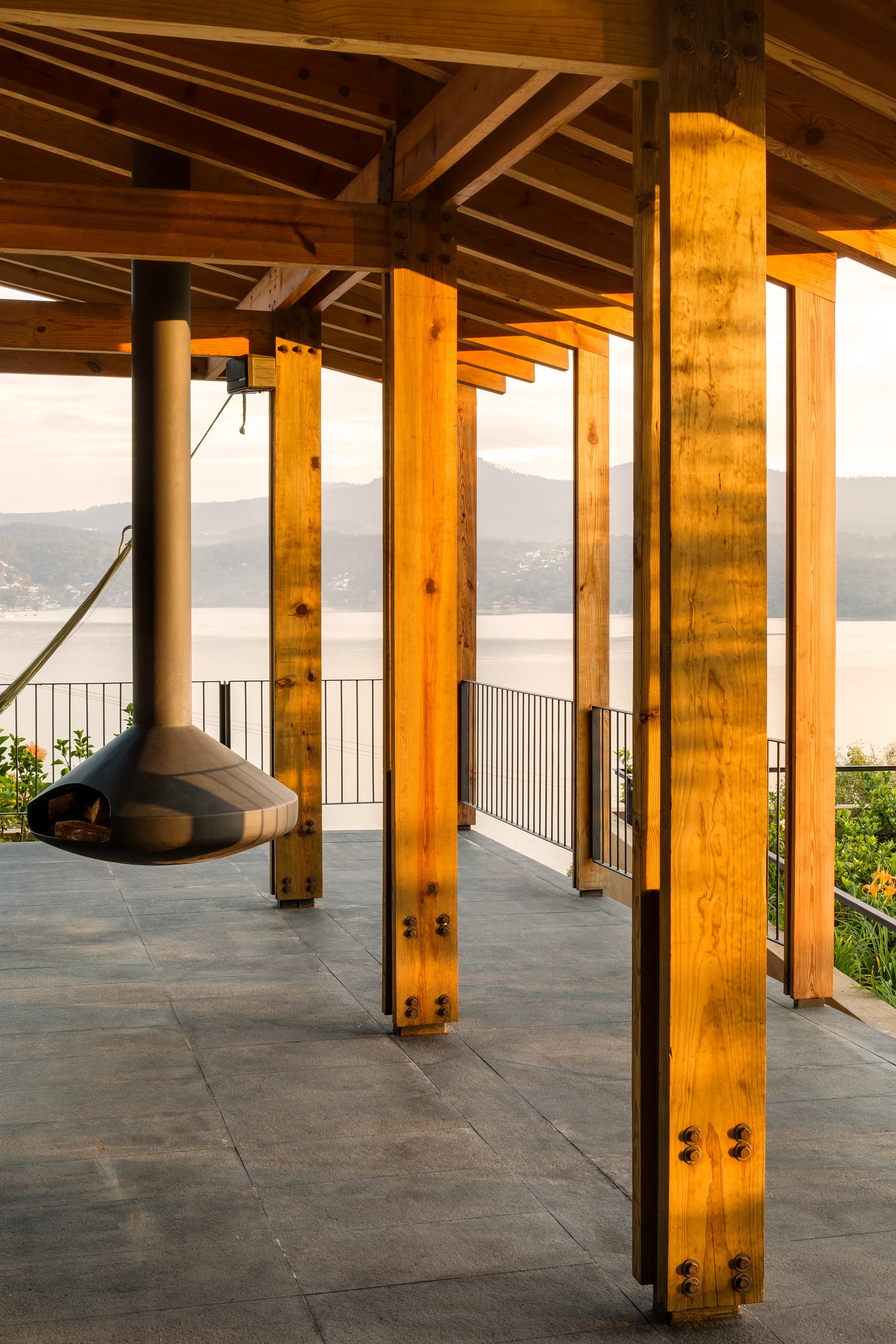 An open-air wooden terrace features stout timber pillars, a dark gray tiled floor, a suspended black fireplace, and a thin metal railing overlooking a lake.