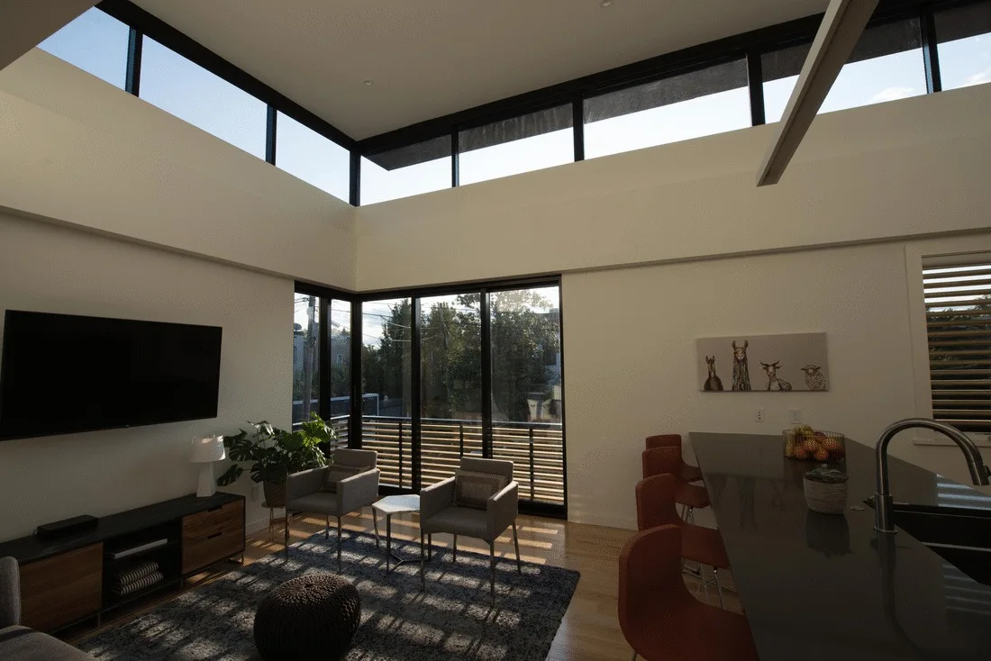 An open-concept living room featuring high clerestory windows, gray armchairs, and a kitchen island with orange stools.