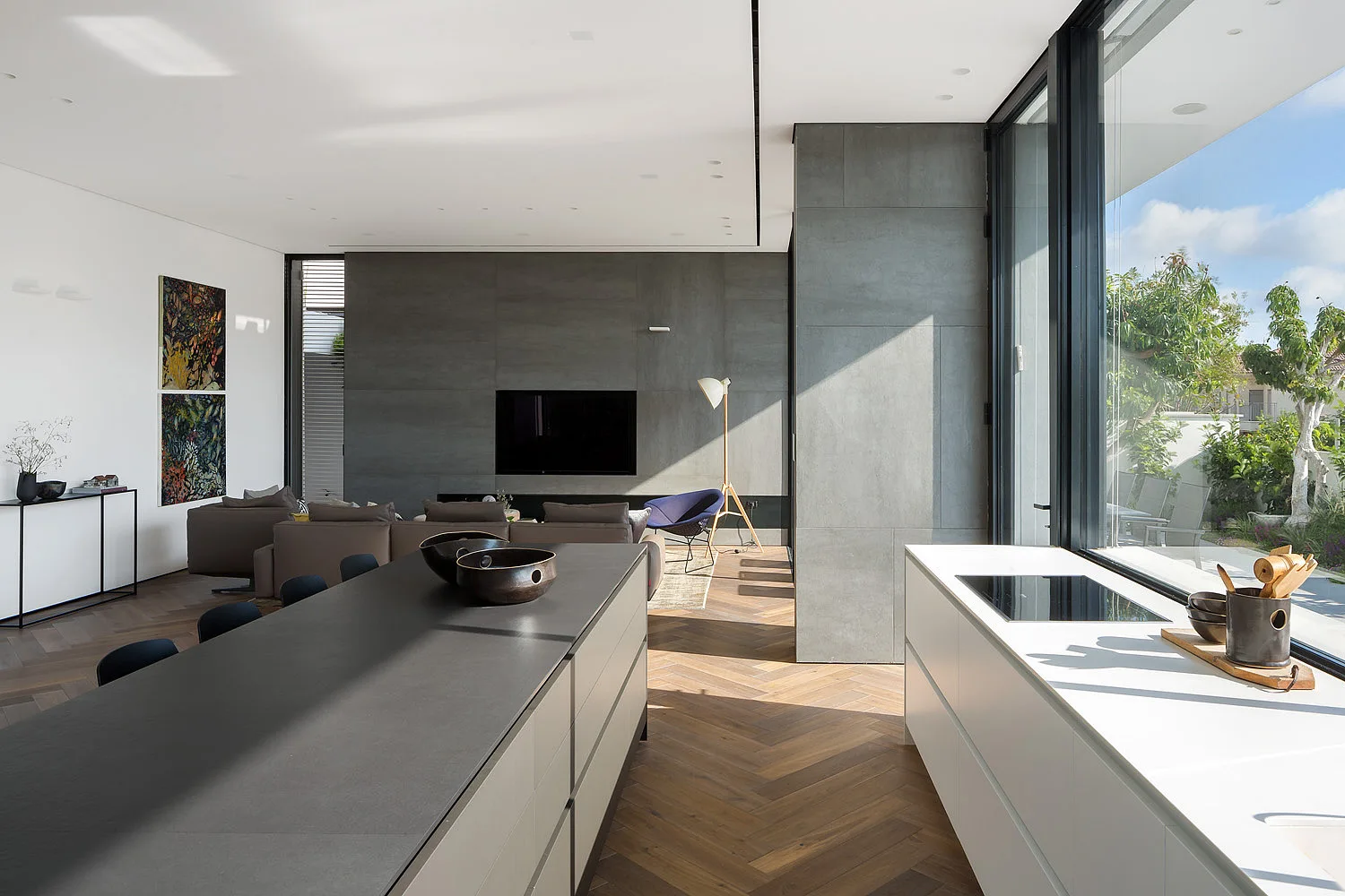 An open-plan kitchen and living space featuring a large dark grey island, herringbone wood floors, and a grey stone-panel feature wall.