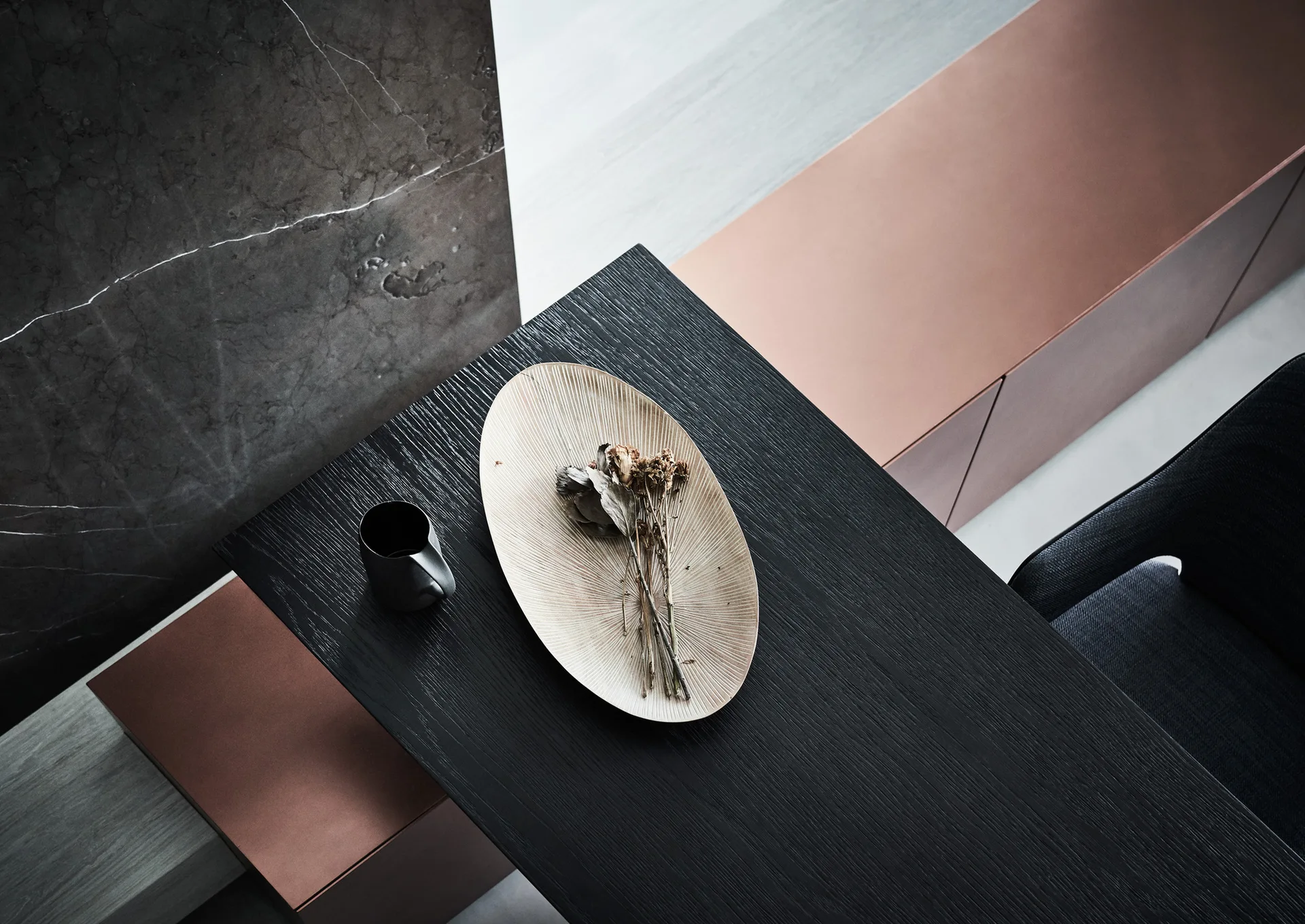 An overhead view of a black wood table with a ribbed tray of dried flowers, set beside copper cabinets and a grey marble wall.