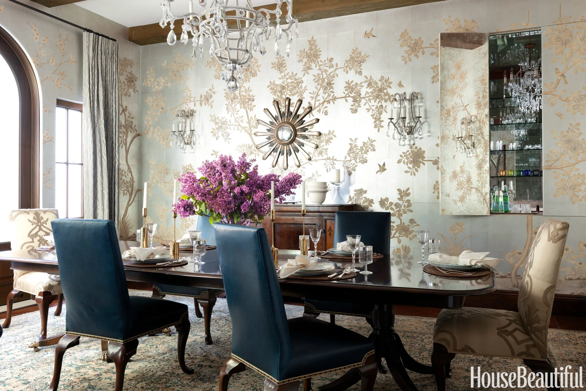 A formal dining room features a dark wood table surrounded by blue leather chairs beneath a crystal chandelier, set against gold floral wallpaper.