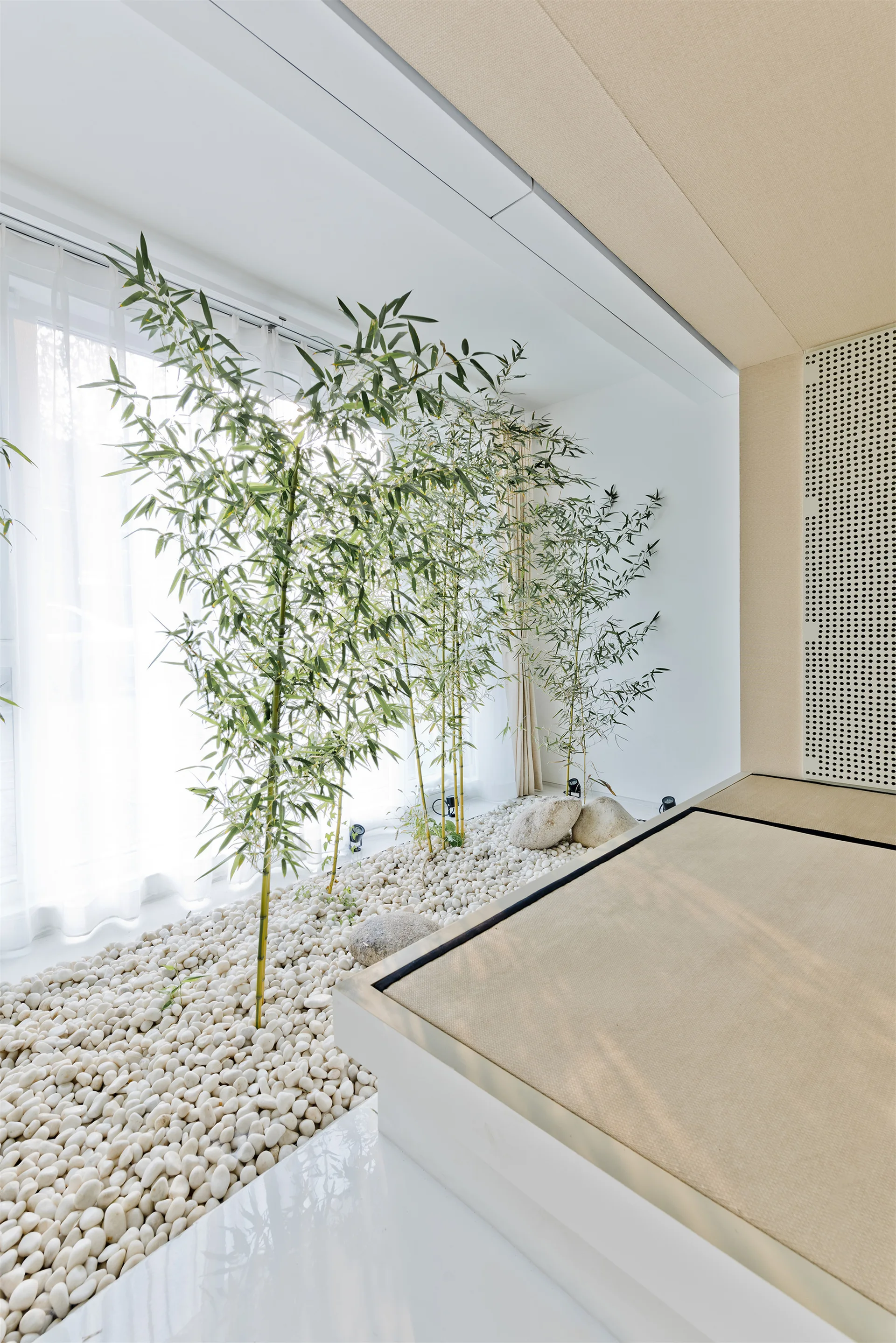 A low platform bed stands next to an indoor garden of green bamboo plants growing out of a bed of smooth white river stones by a window.