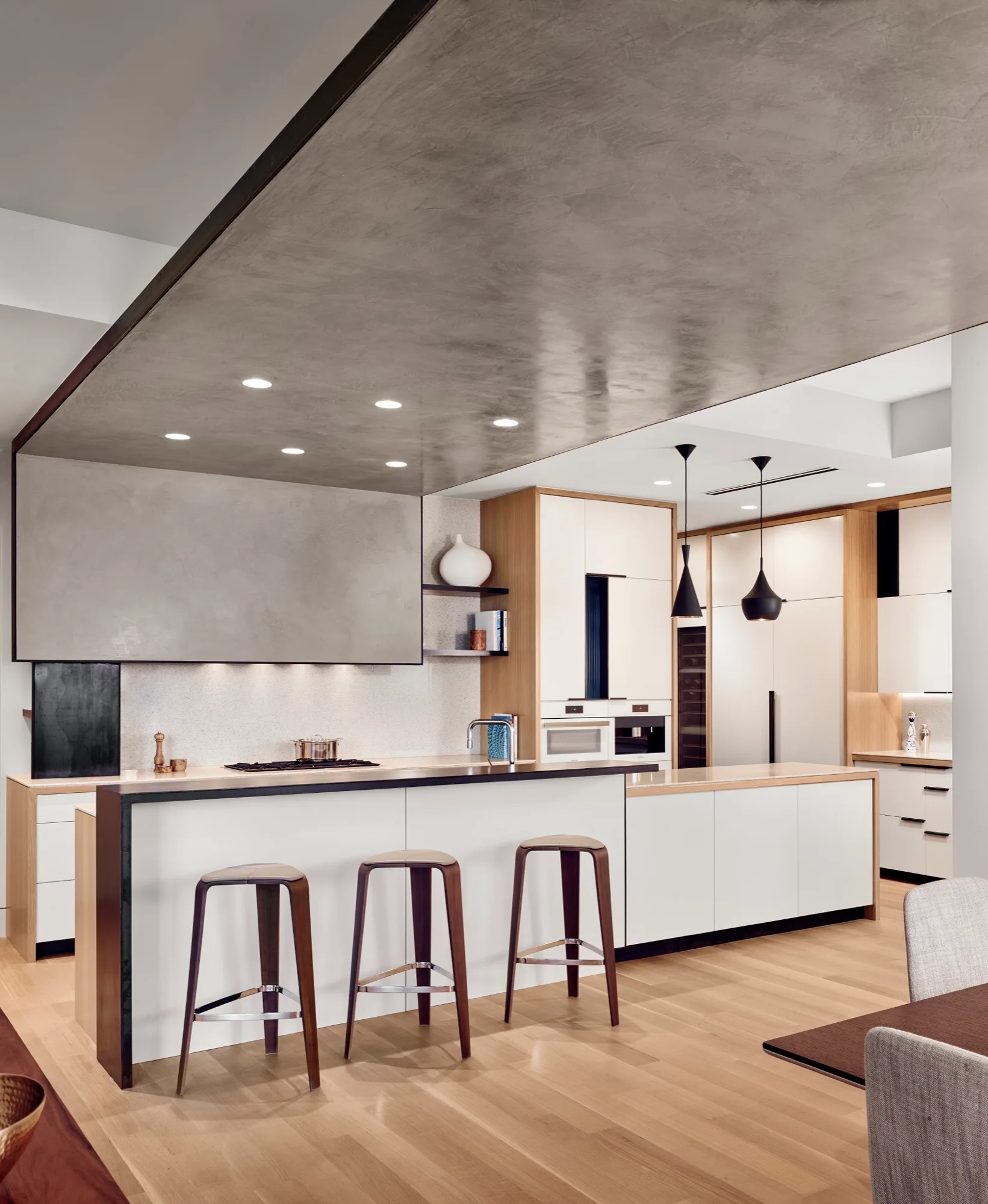 A modern kitchen features a white island with three wood bar stools under a large grey ceiling soffit, with matching integrated cabinetry behind.