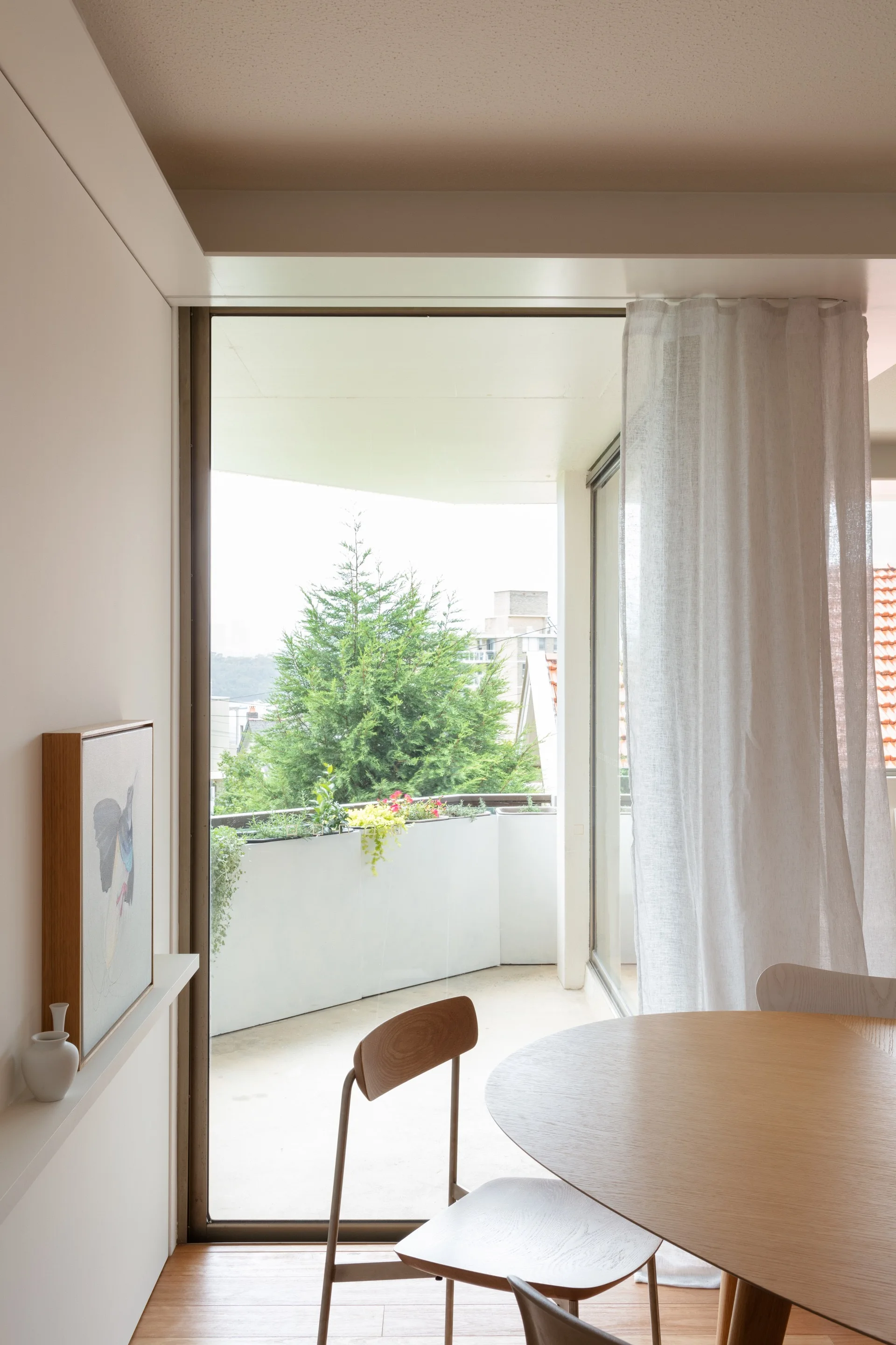A light wood round dining table and minimalist chairs sit next to a large glass door leading to a balcony with green plants.