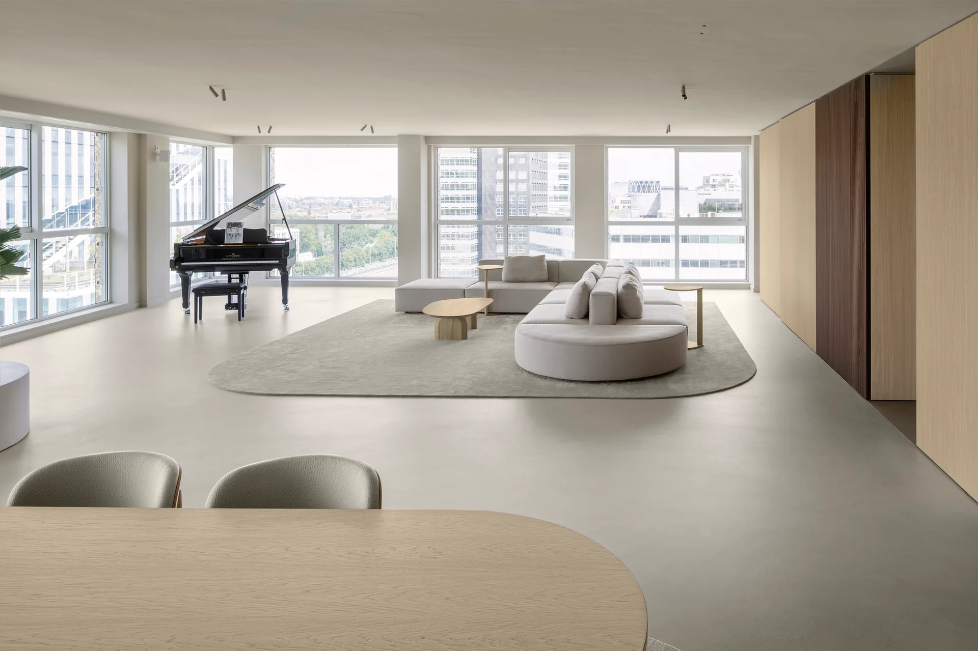 A spacious minimalist living room features a cream modular sectional sofa on an oval rug, a black grand piano, and floor-to-ceiling windows.
