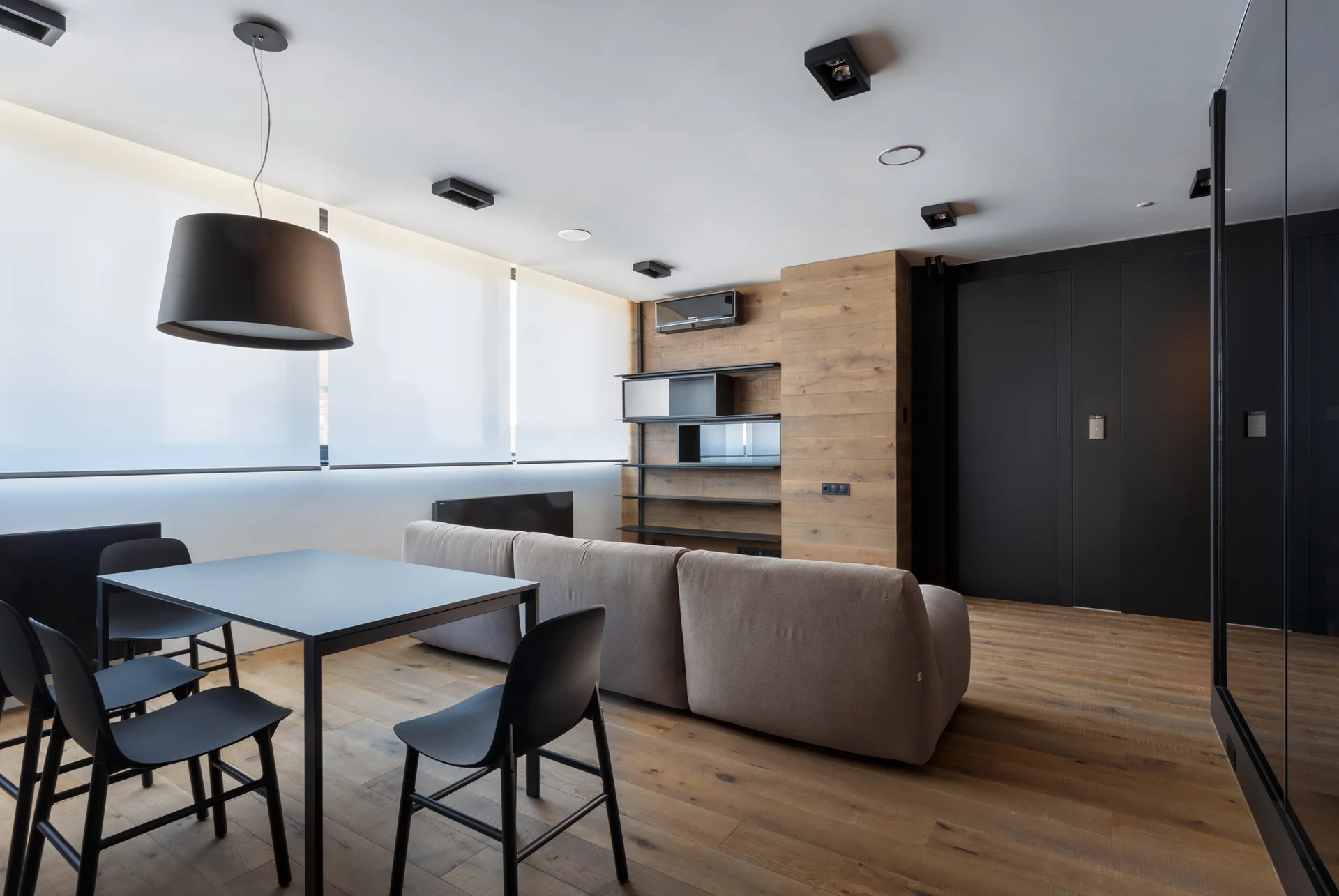 A modern open-plan living and dining room featuring a beige sofa, black dining set, and a wood-paneled accent wall.