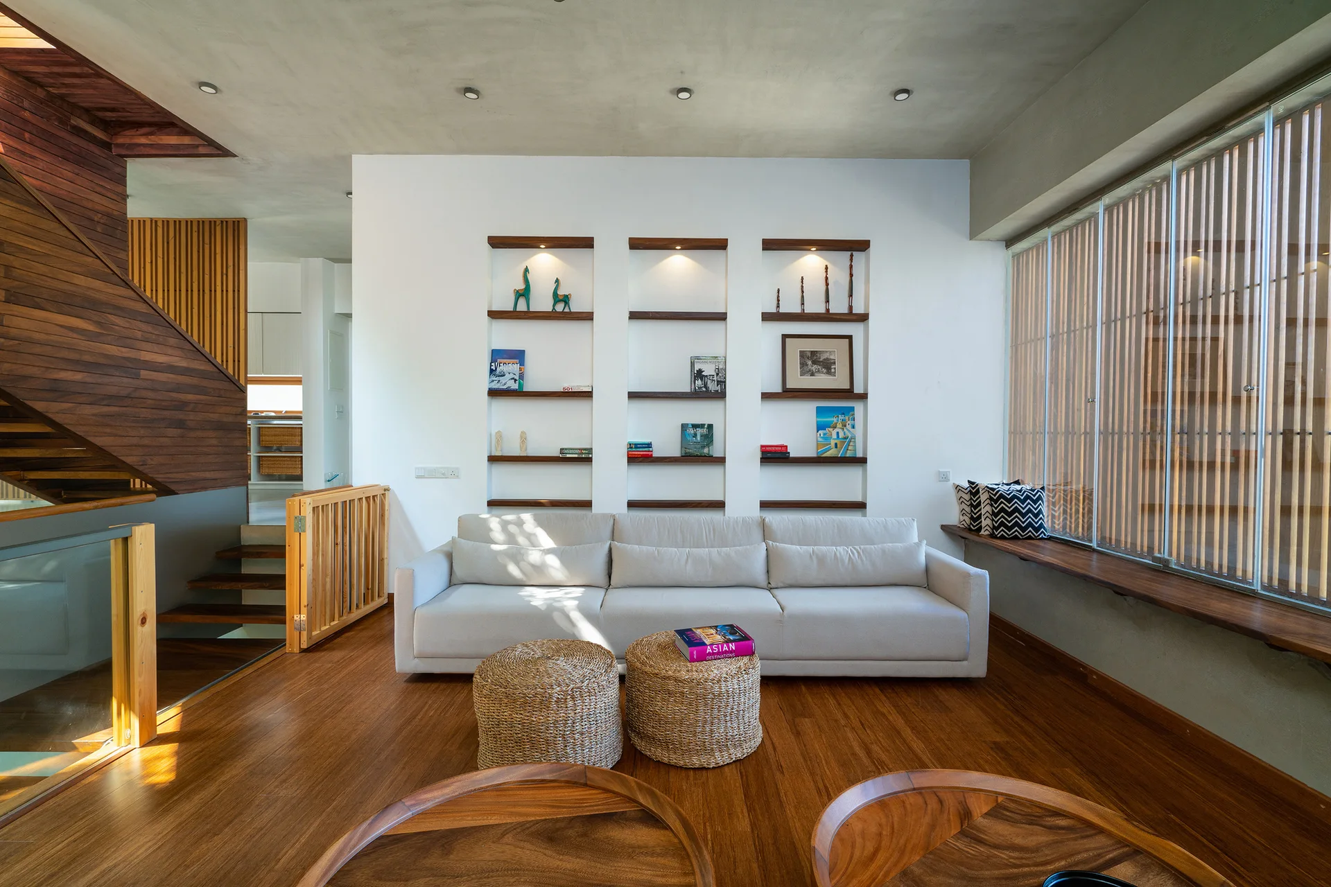 A modern living room features a cream sofa, two woven ottomans, and three recessed wall niches with wood shelves beneath an industrial concrete ceiling.