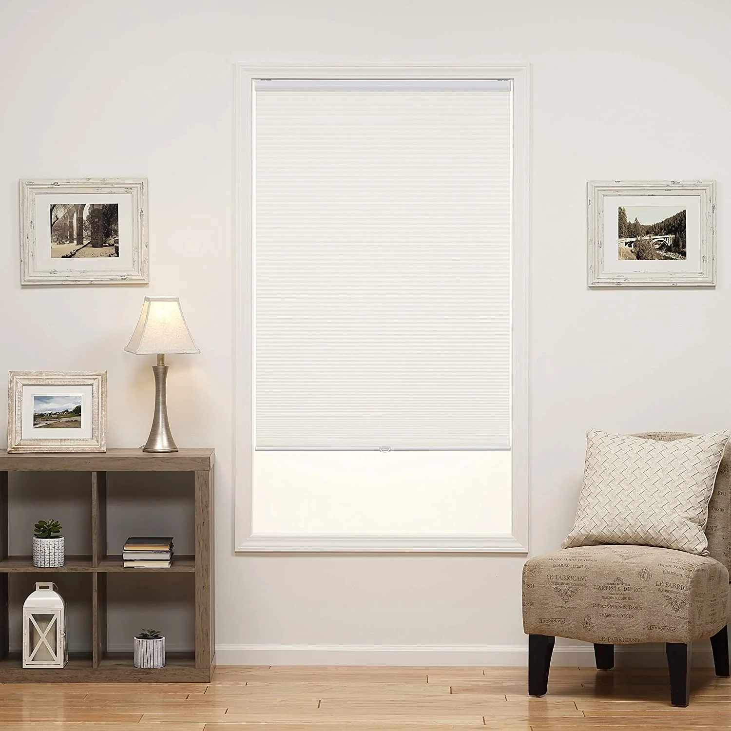 A white pleated cellular shade covers a window flanked by framed art prints above an open wooden shelving unit and a script fabric accent chair.