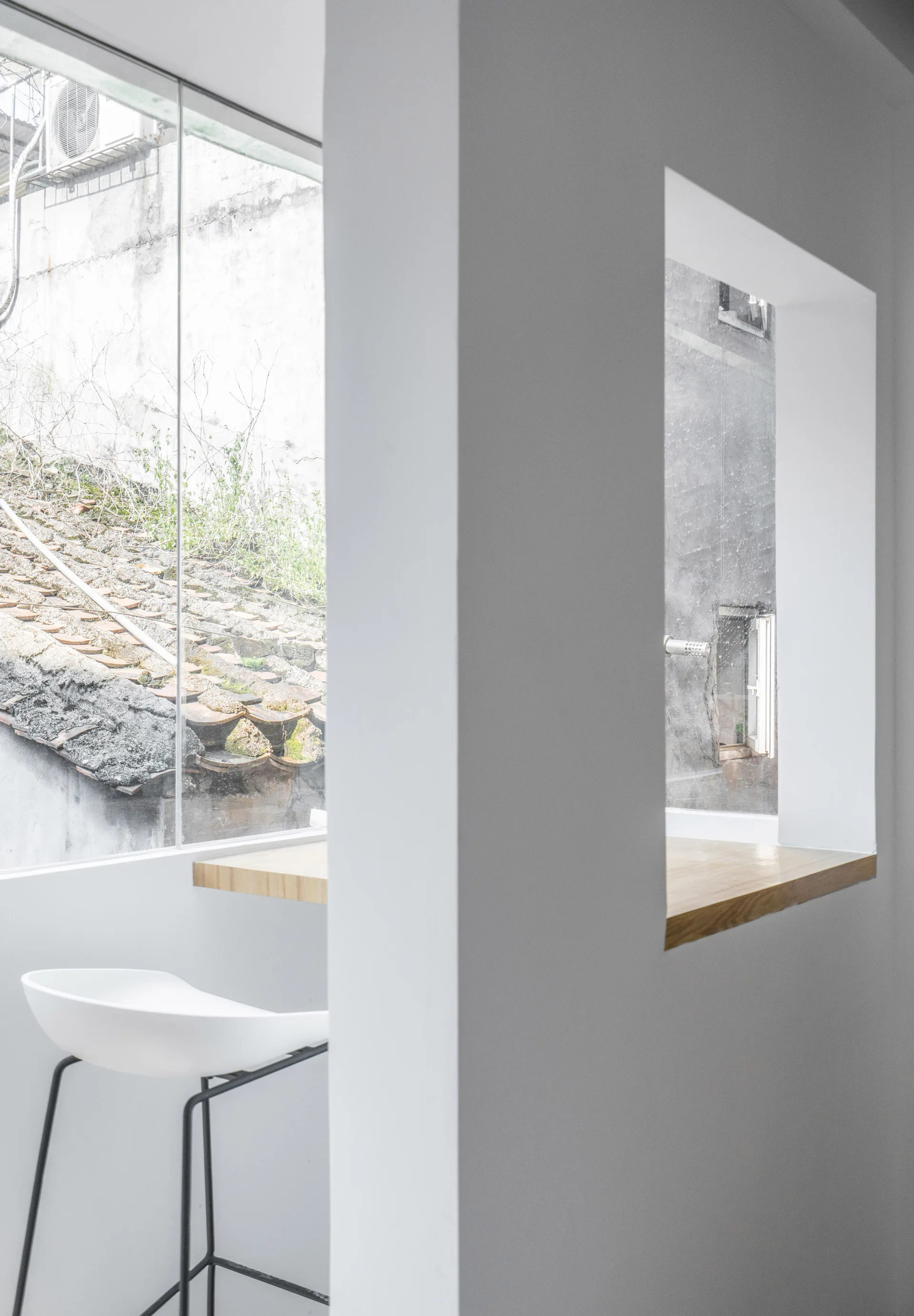 A minimalist interior features a white bar stool next to a wooden window ledge overlooking an old tiled roofscape.
