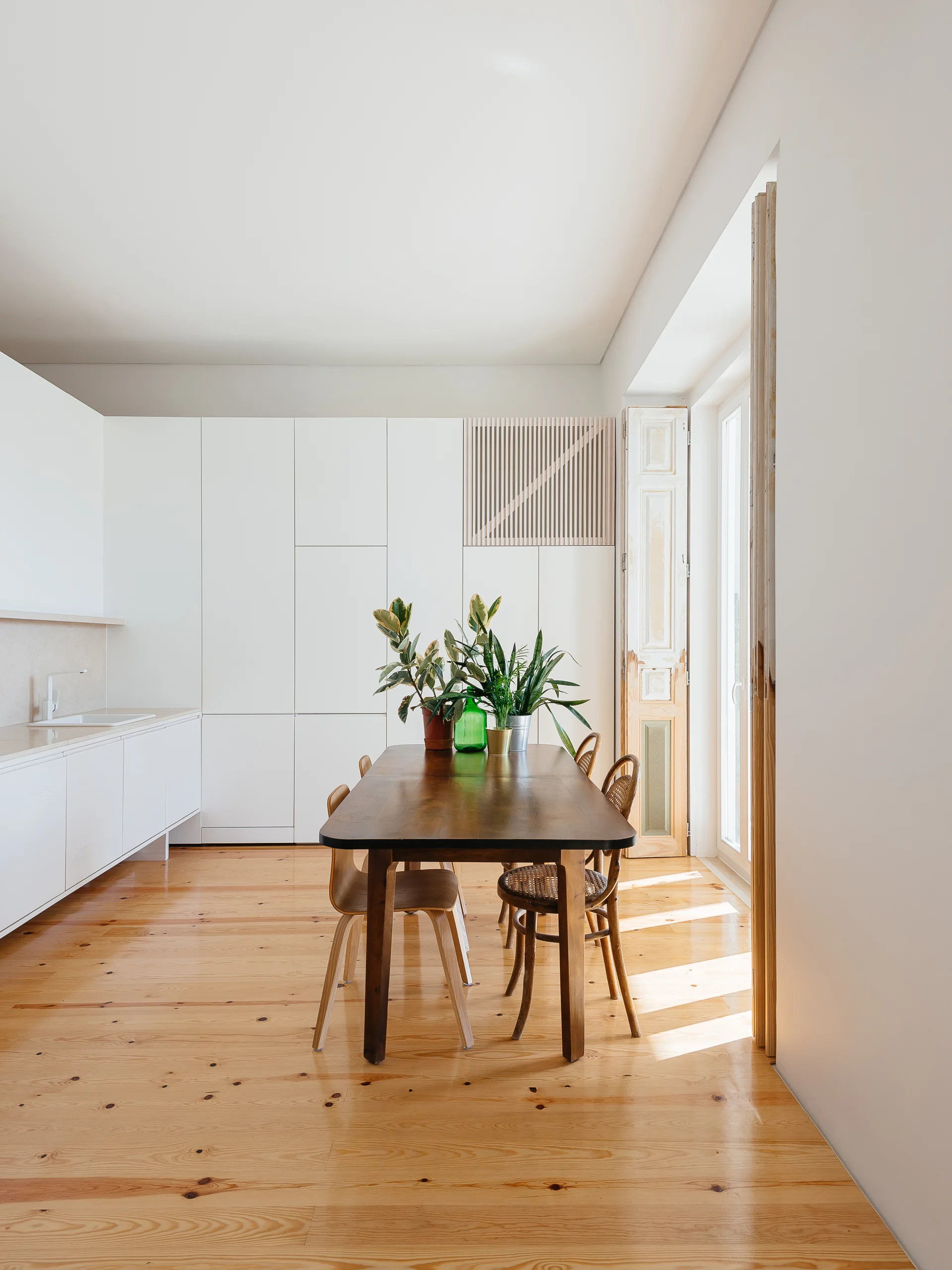A dark wood dining table and mismatched wooden chairs sit on light pine floors in front of seamless white kitchen cabinets.