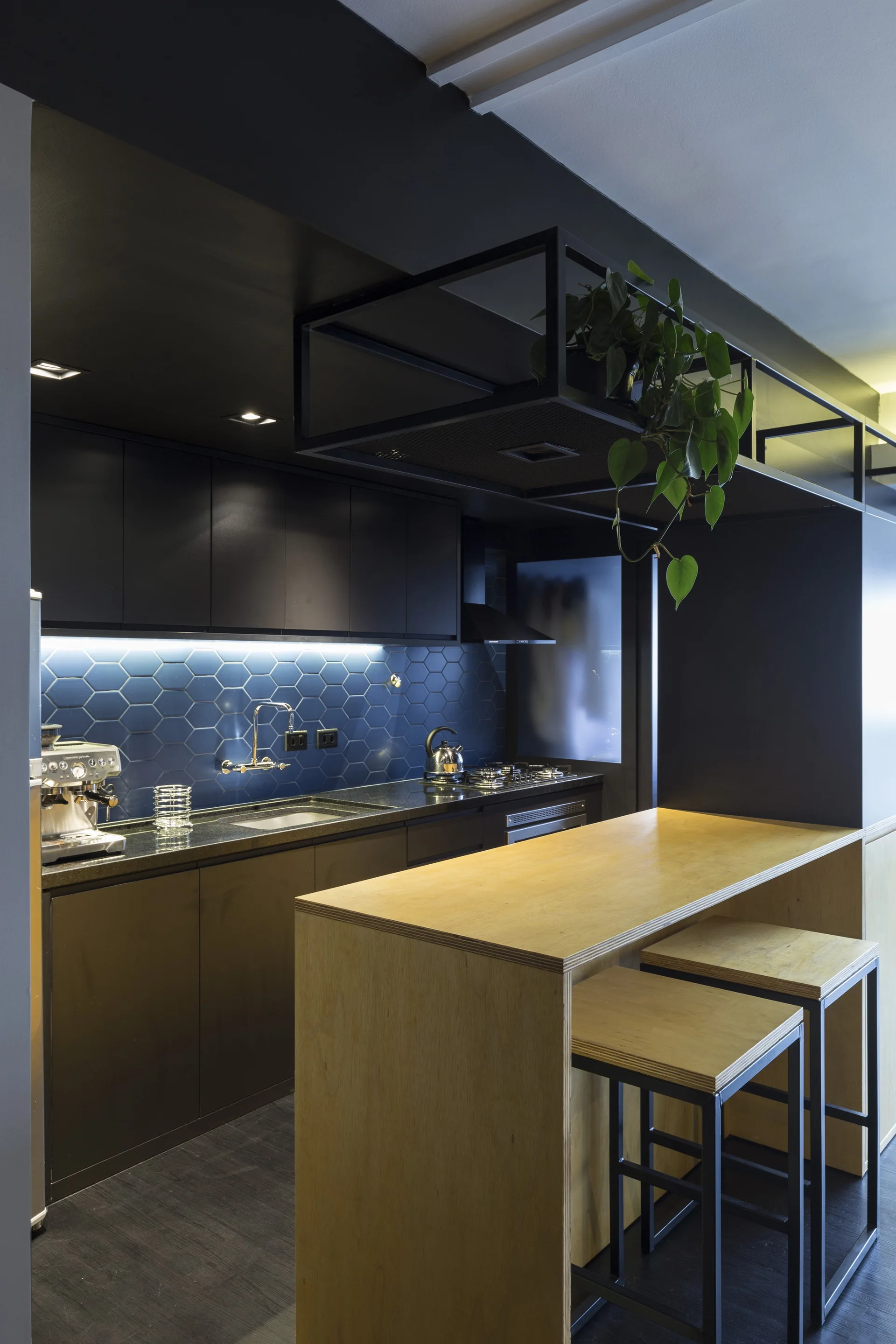 A modern kitchen featuring matte black cabinets, a blue hexagonal tile backsplash with under-cabinet lighting, and a light wood island with stools.