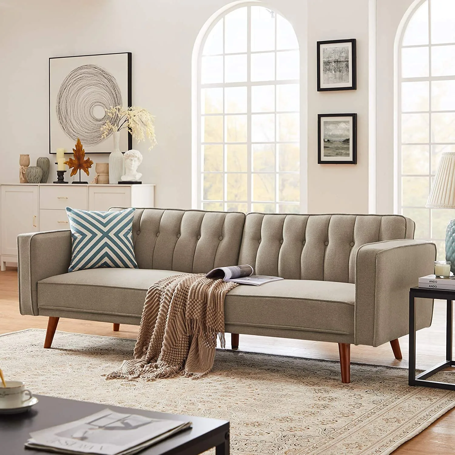 A grey tufted sofa with a textured throw blanket sits on a patterned rug in front of large arched windows in a bright living room.