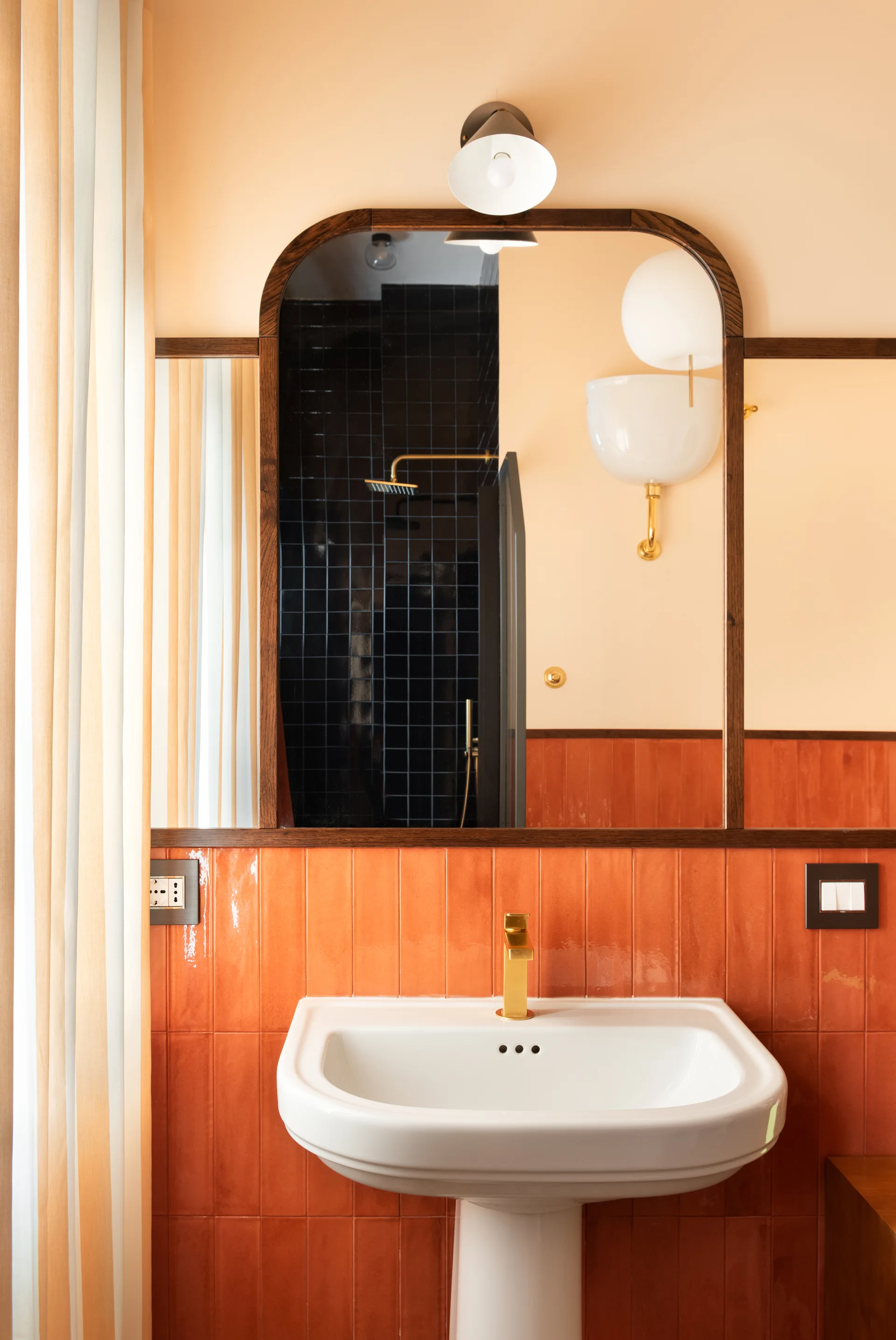 A white pedestal sink with a gold faucet sits against glossy orange tiles below an arched wood-framed mirror in a warm-toned bathroom.