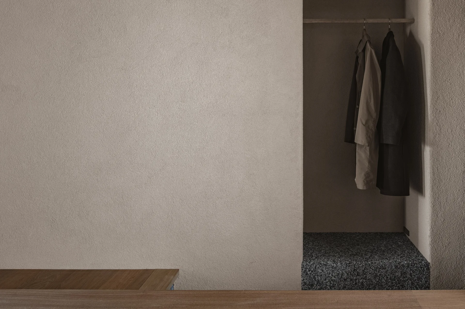 A recessed open closet niche built into a textured beige plaster wall contains dark coats hanging on a simple wooden rod above a pebbled stone base.