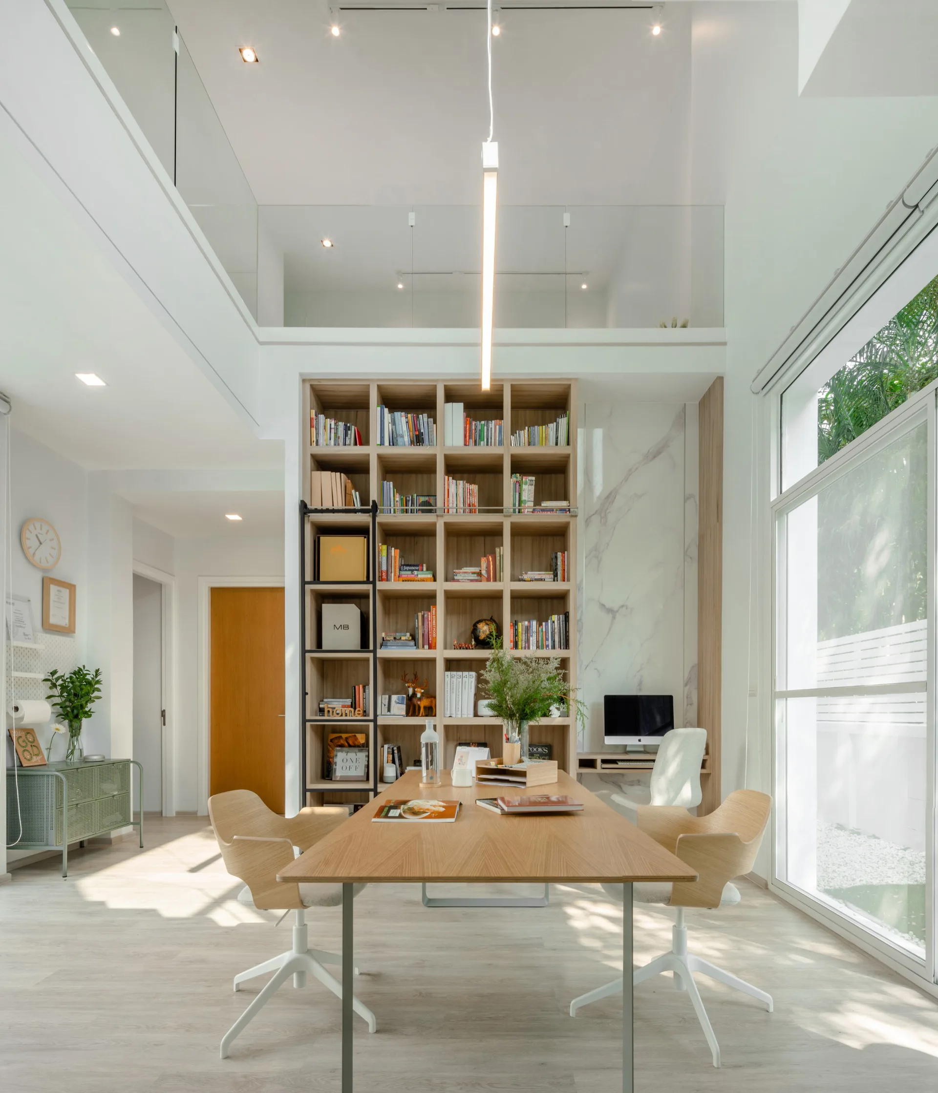 A bright double-height home office featuring a large light wood desk, a tall built-in wooden bookshelf with a black ladder, and floor-to-ceiling windows.