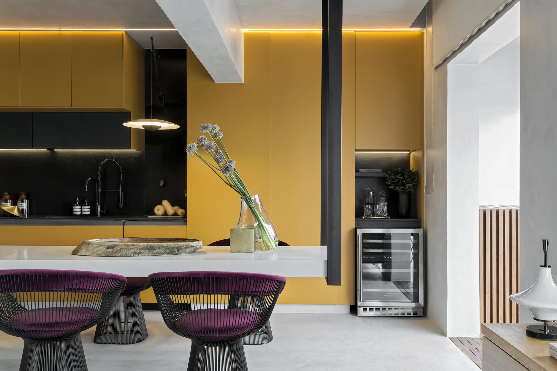 A modern kitchen featuring yellow cabinetry, a white stone island, and black wire dining chairs with purple velvet cushions.