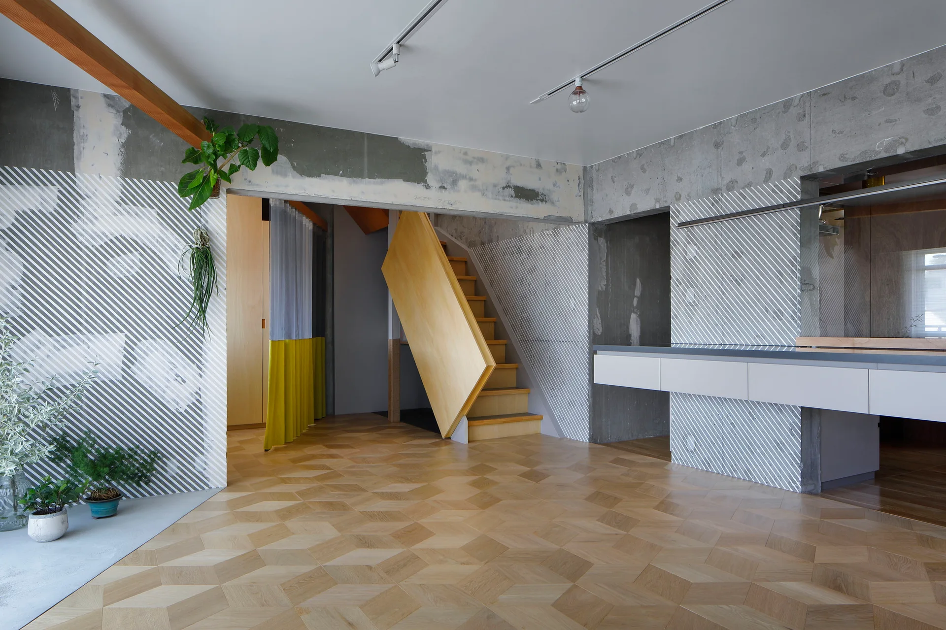 A minimalist room features concrete walls painted with diagonal white stripes, geometric wood parquet floors, and an angled wooden staircase.