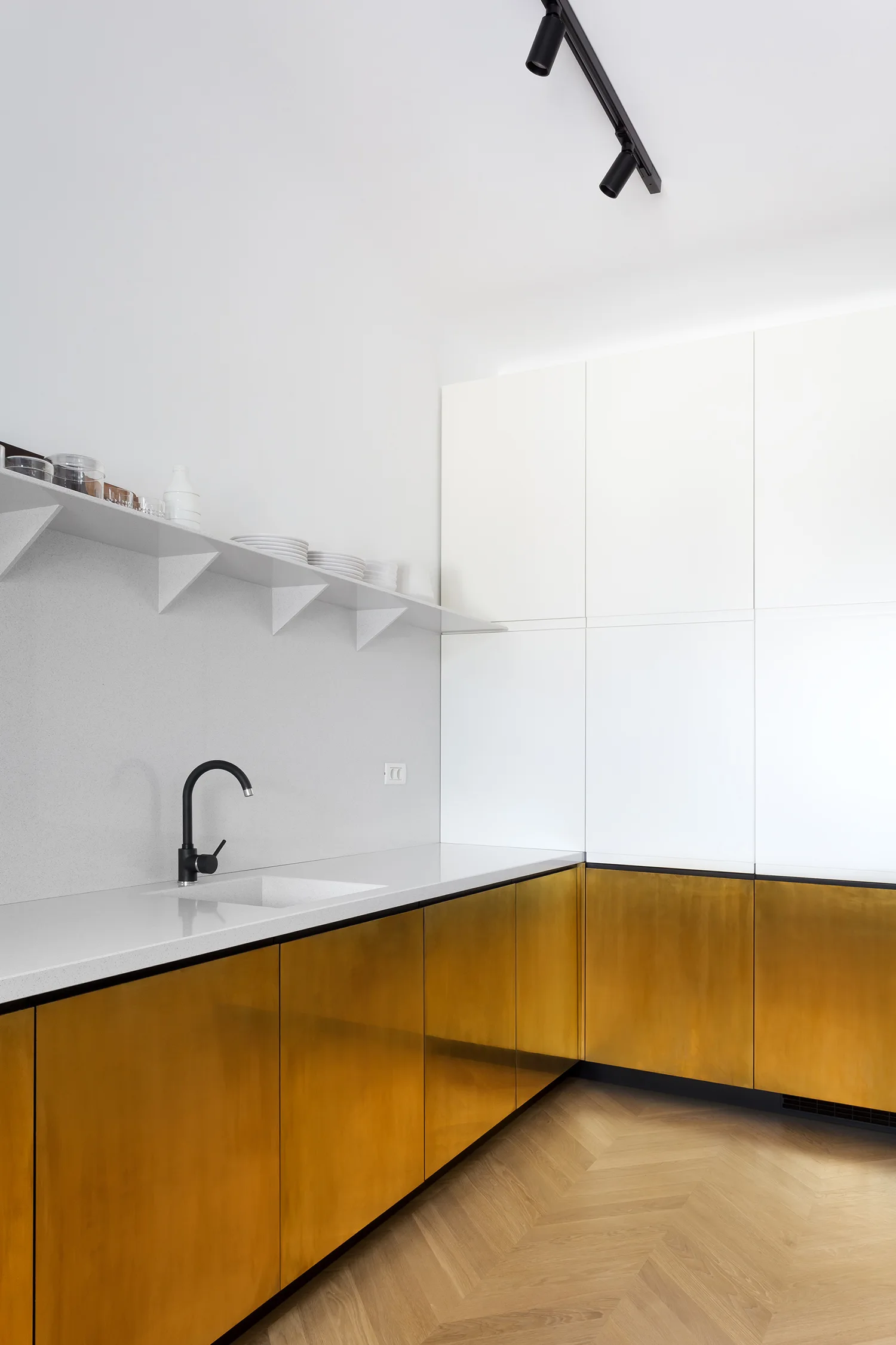 A modern kitchen featuring metallic brass lower cabinets, white upper cabinets, a white countertop with a black faucet, and herringbone wood floors.