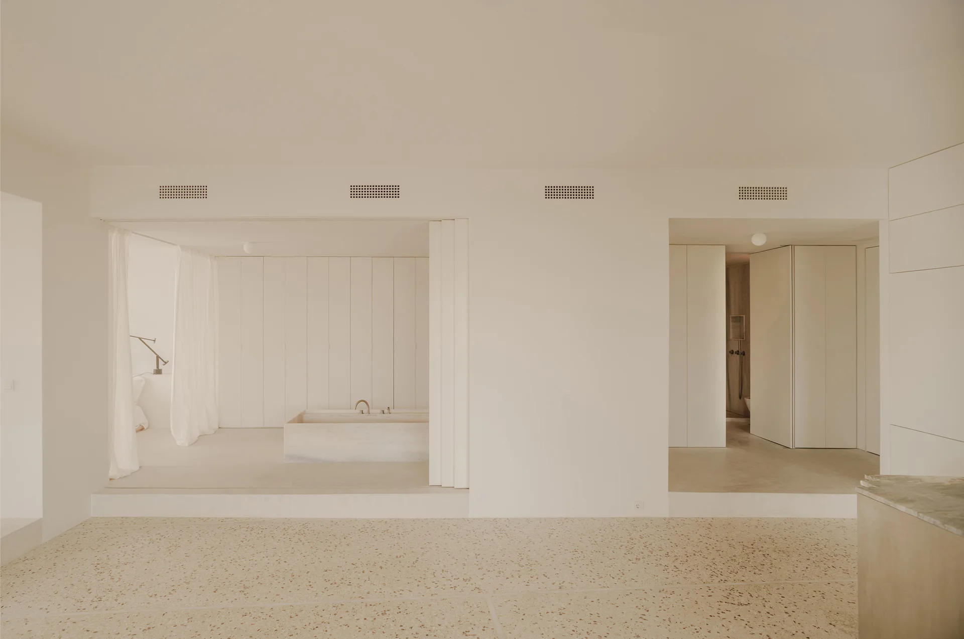 A minimalist beige bathroom featuring a stone rectangular bathtub on a raised platform, floor-to-ceiling wall panels, and terrazzo flooring.