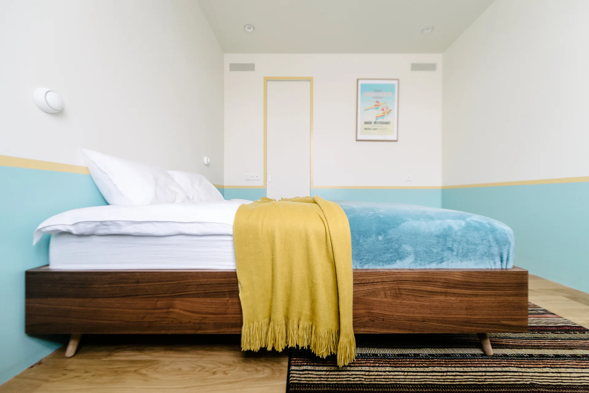A minimalist bedroom features a wooden platform bed with a yellow throw blanket against a two-tone blue and white painted wall.