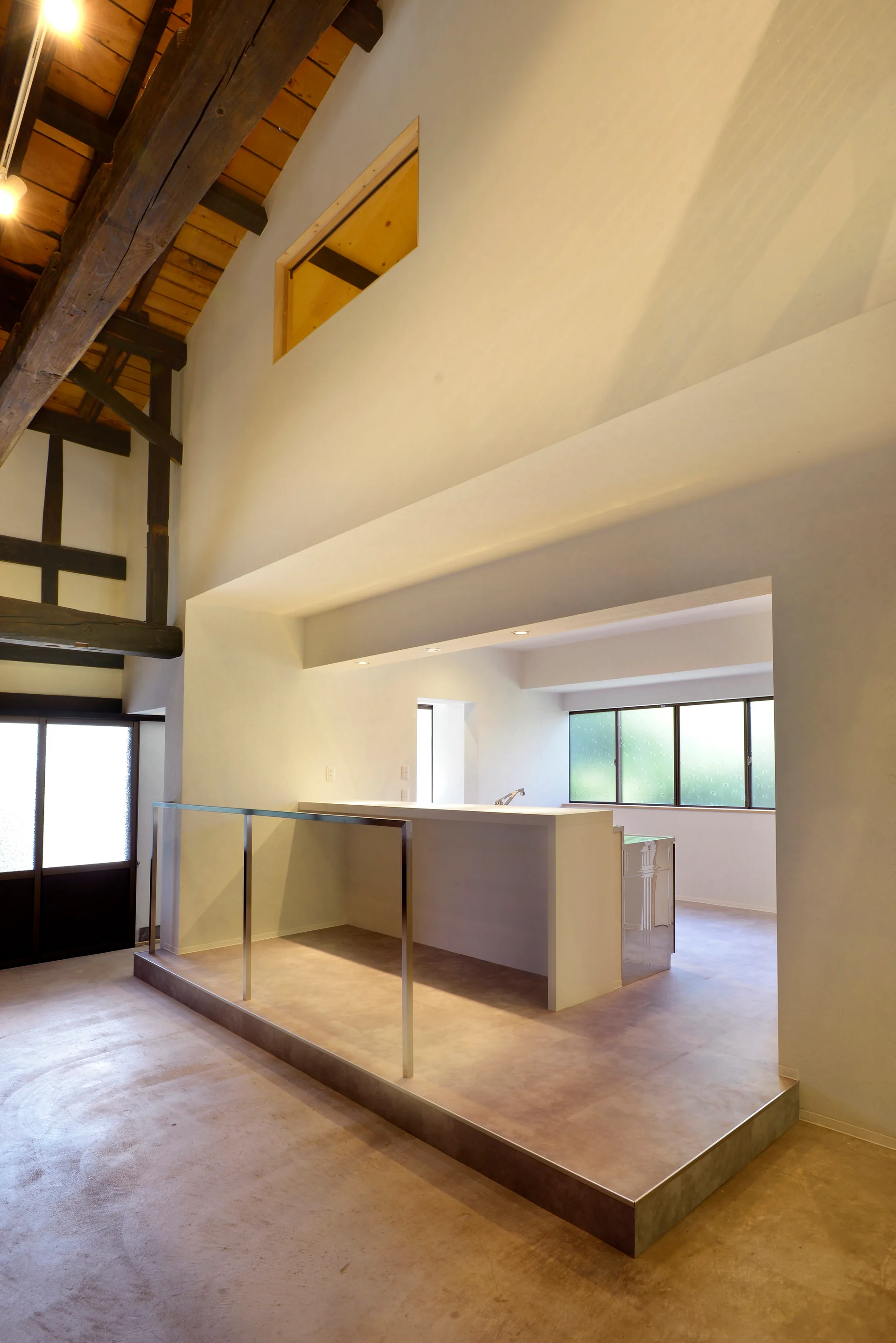 A minimalist kitchen on a raised platform features a white island beneath a vaulted ceiling with dark exposed timber beams.