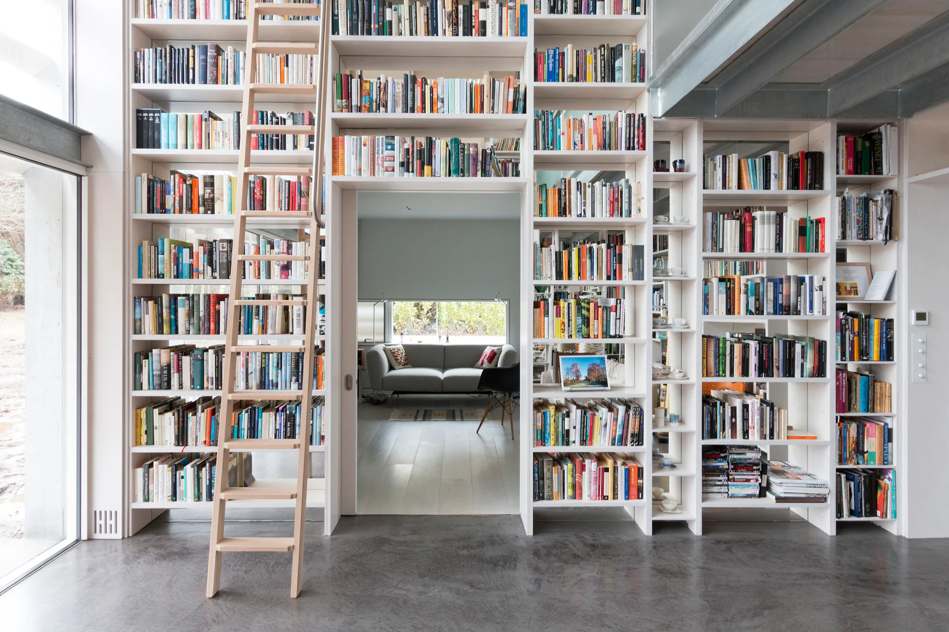 A tall white built-in bookshelf surrounds a doorway, featuring a light wood rolling ladder and a view of a grey sofa in the next room.