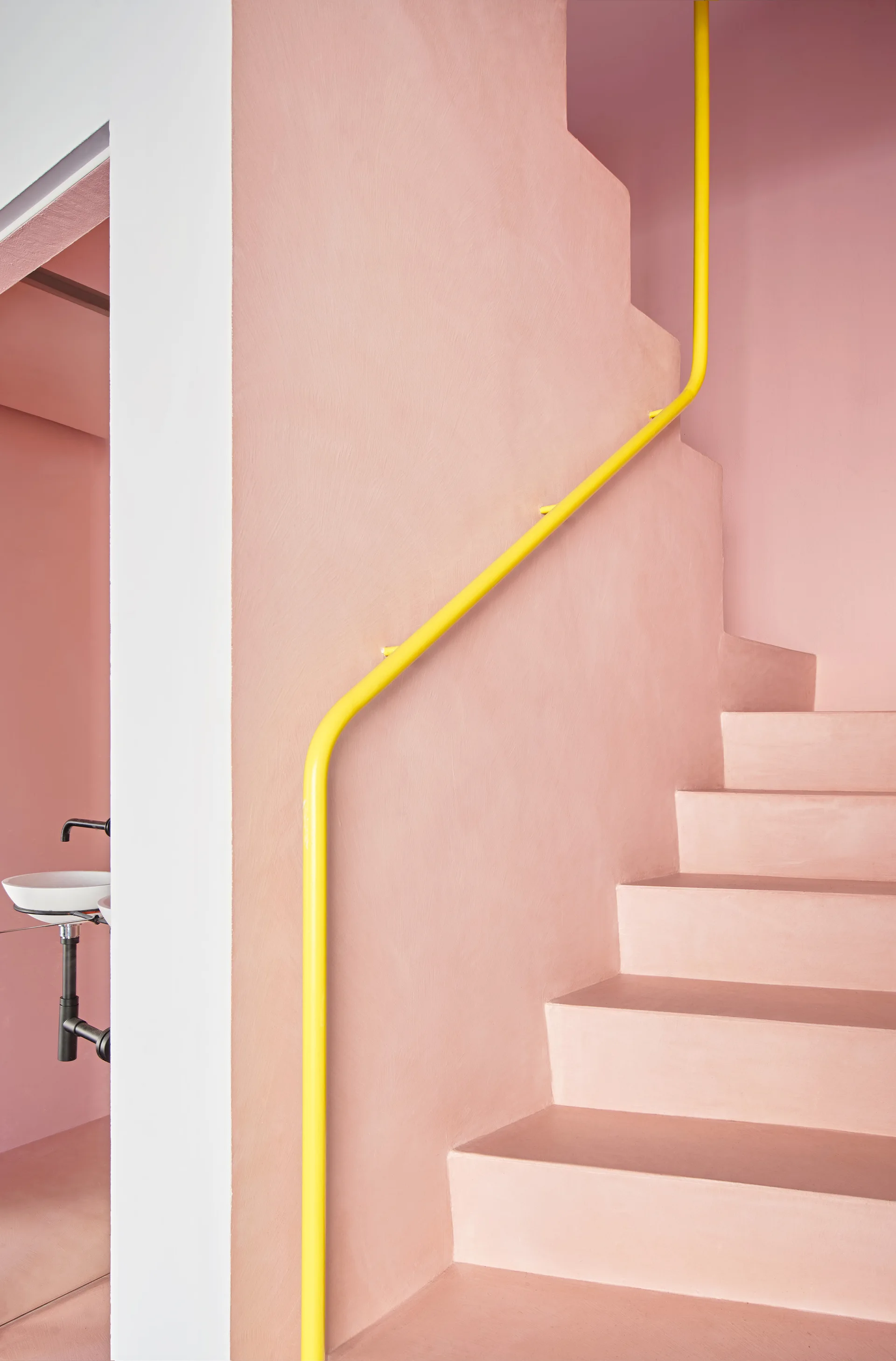 A minimalist staircase painted entirely in matte pink features a bright yellow metal handrail and a glimpse of an adjacent white washbasin.