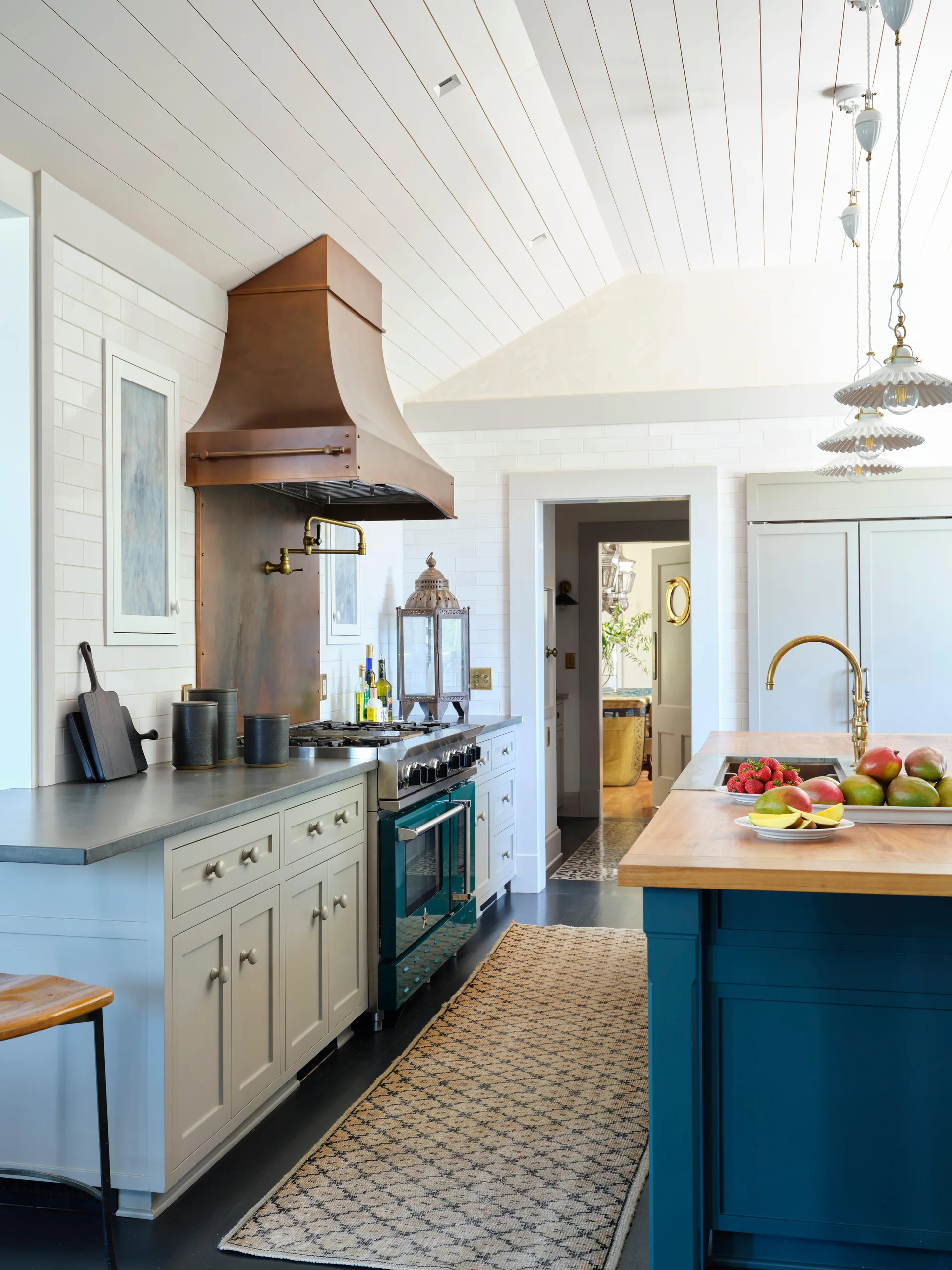 A kitchen features a copper range hood, a dark green stove, light grey lower cabinets, and a blue island with a wood countertop.
