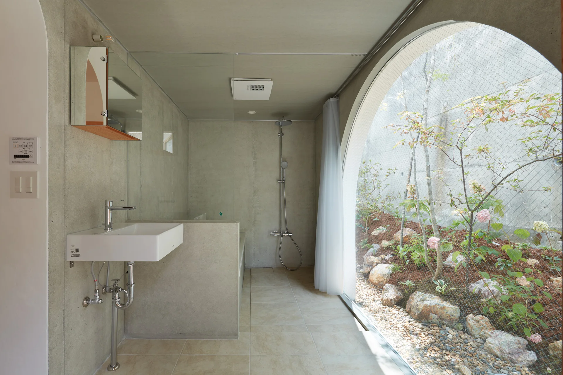 A minimalist gray concrete bathroom features a wall-mounted white sink and a large arched glass window looking out onto a small private garden.