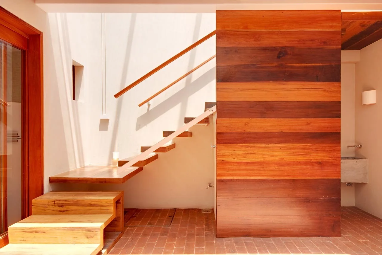 A minimalist floating wood staircase features a glass railing next to a horizontal wood paneled wall and a terracotta brick floor.