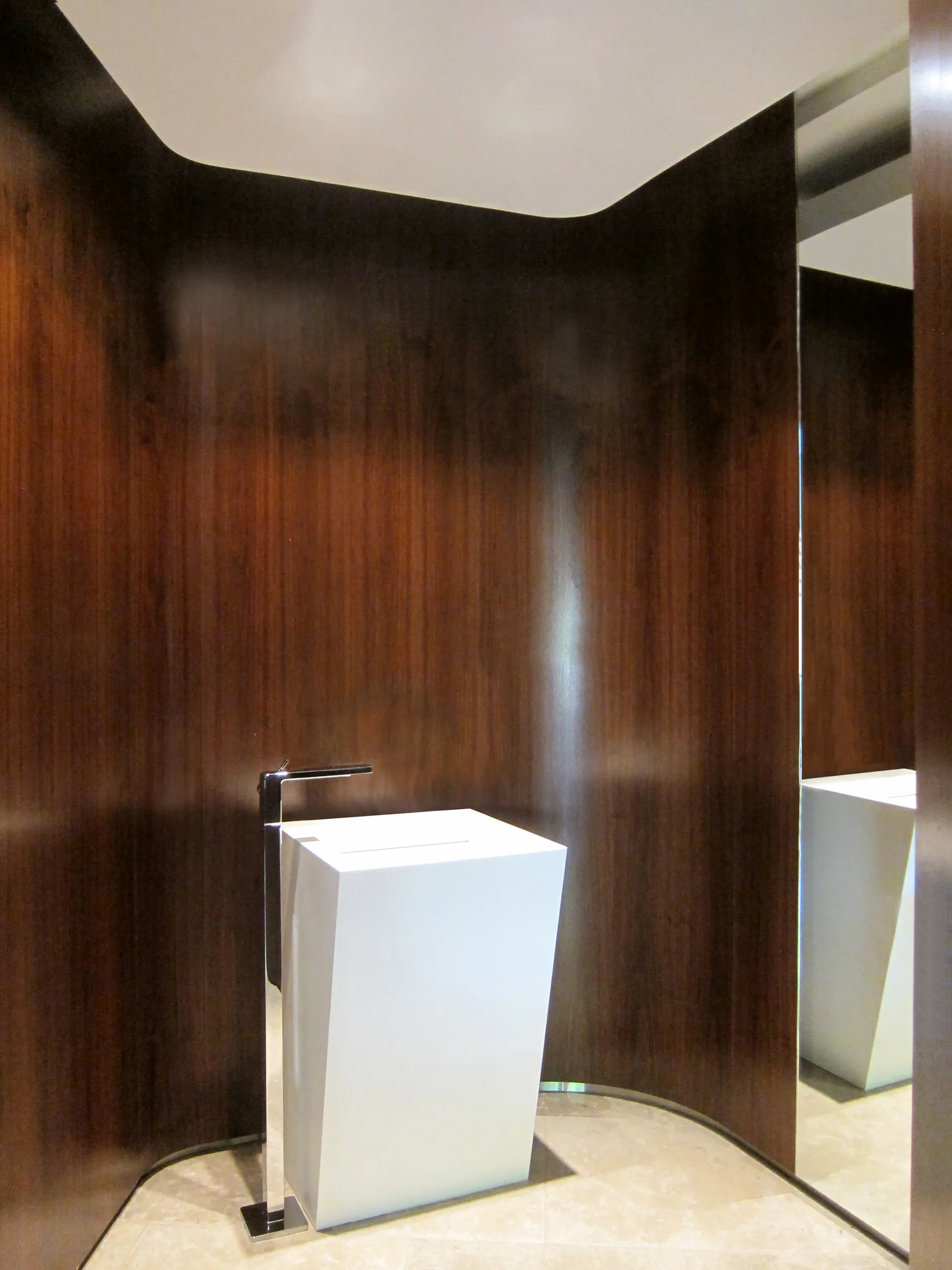 A modern white pedestal sink with a chrome faucet sits in a bathroom nook featuring curved dark wood paneling and a large mirror.