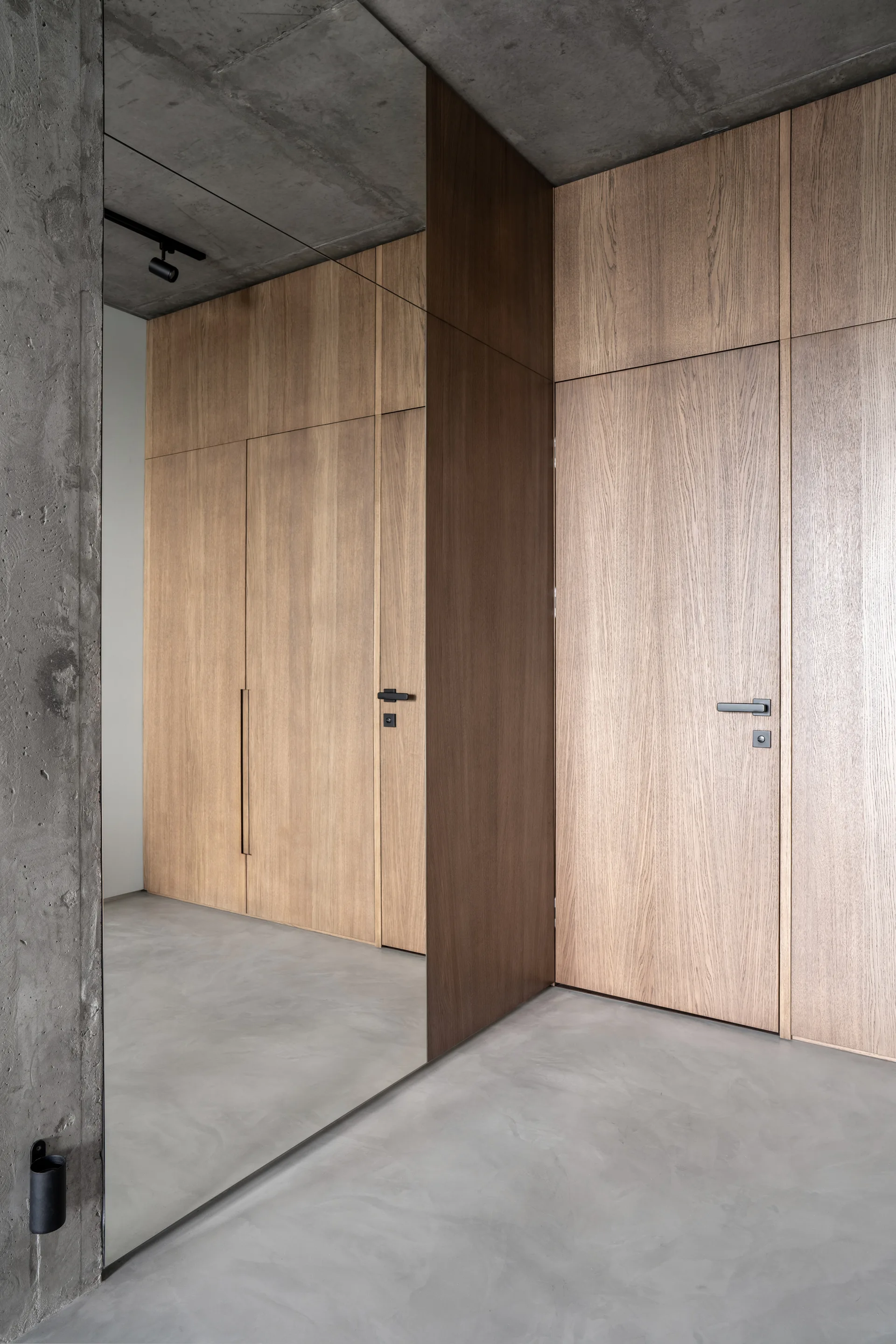 Floor-to-ceiling light wood cabinetry and a large vertical mirror in a hallway with raw concrete floors and ceiling.