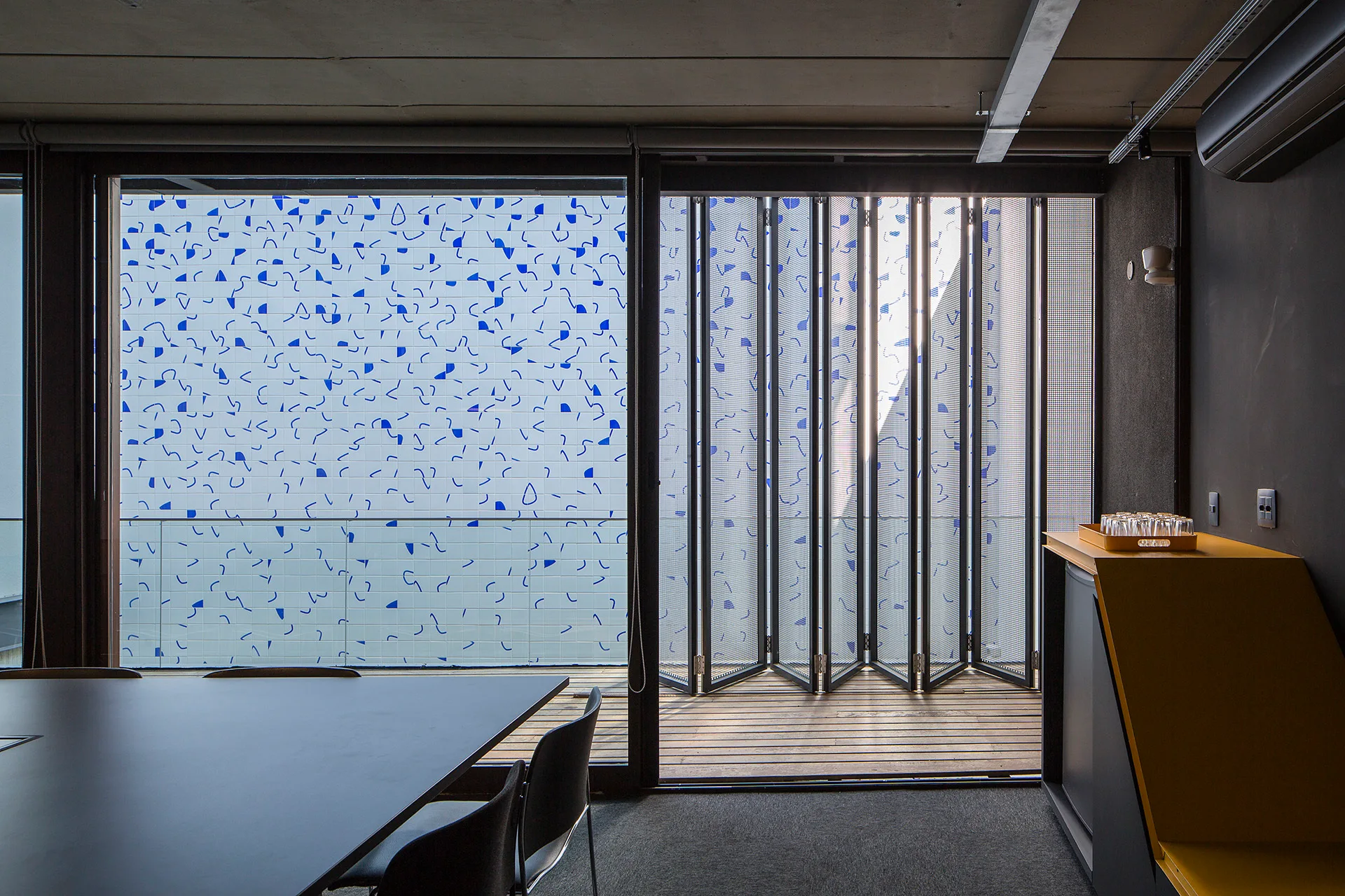 A modern dark grey meeting room features a concrete ceiling, folding glass doors, and a balcony with blue and white patterned wall tiles.