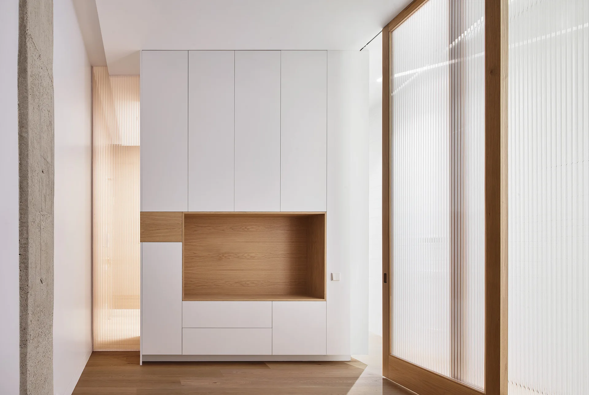 A modern minimalist hallway featuring a built-in white storage cabinet with an oak niche next to a fluted glass sliding door.