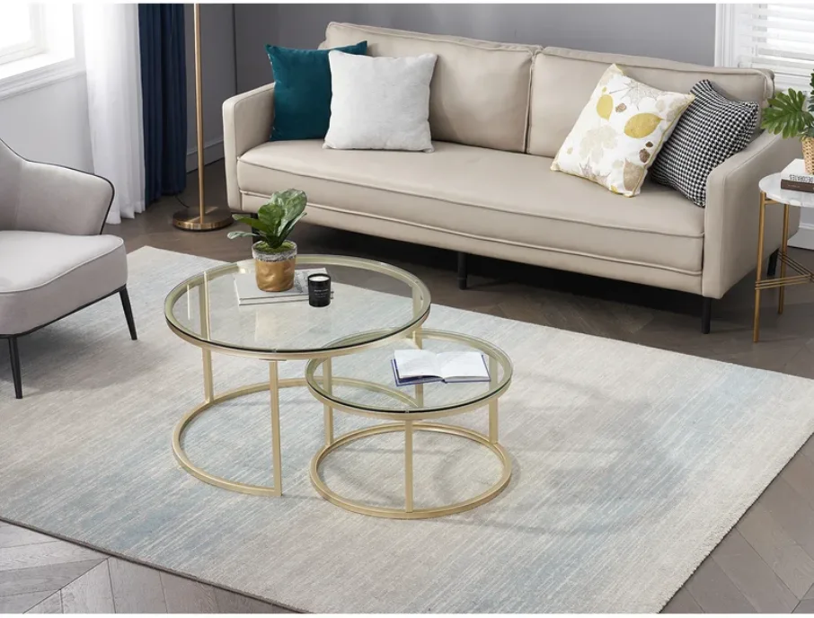 Two round glass coffee tables with gold legs sit on a grey rug in front of a beige sofa on a herringbone wood floor.