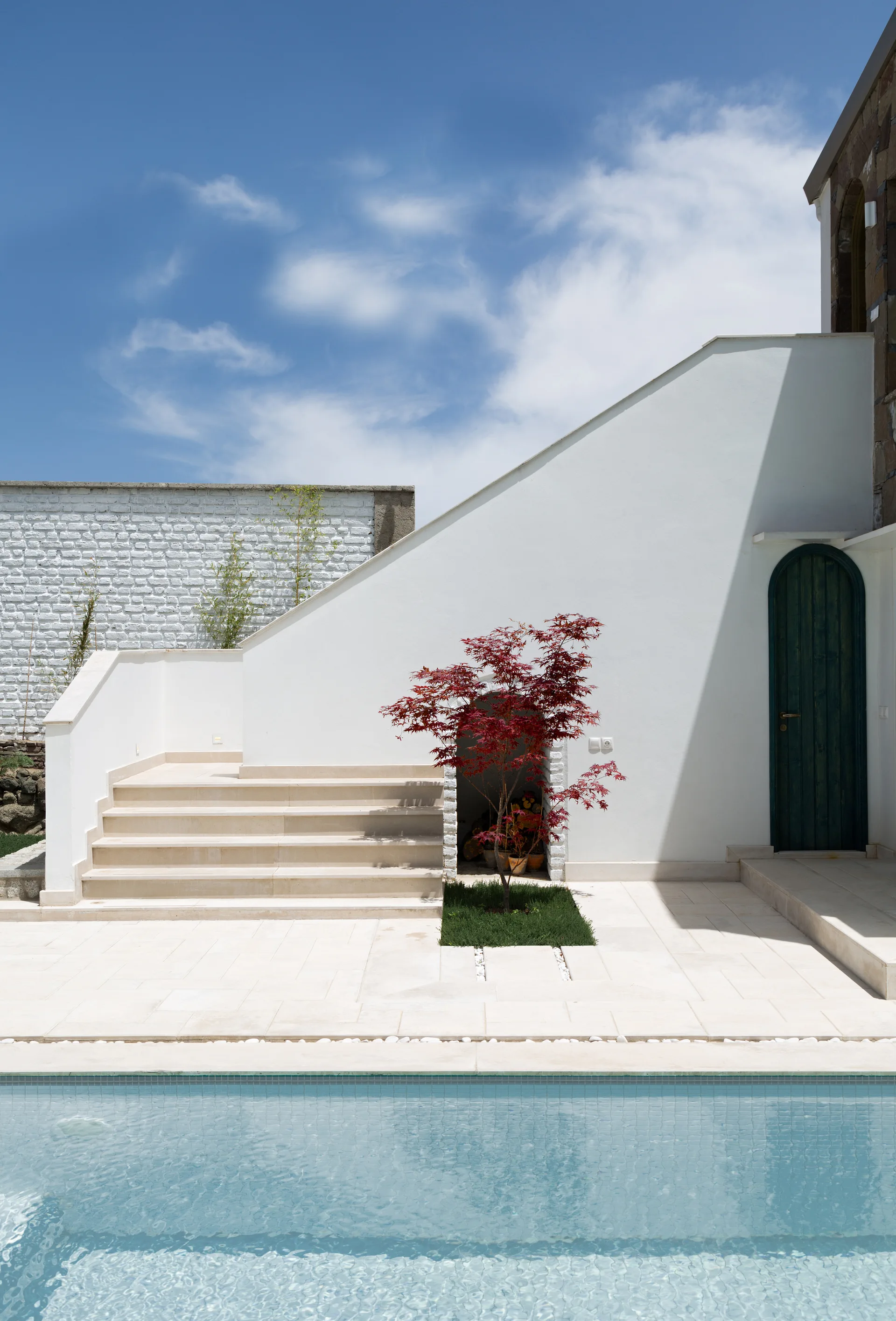 An outdoor swimming pool fronts a white plaster patio with beige stone stairs, an arched green door, and a small red Japanese maple tree.