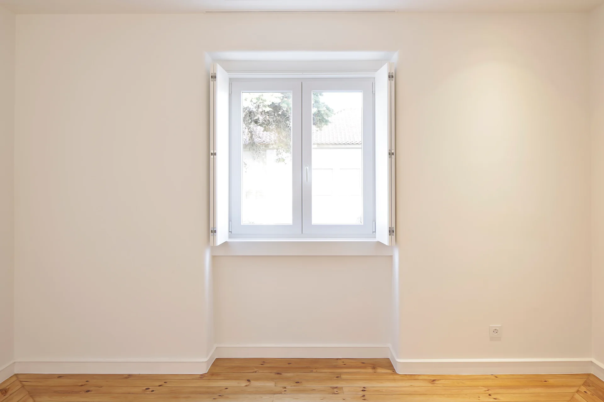 An empty room featuring smooth white walls, natural pine hardwood flooring, and a central window with open white wooden shutters.